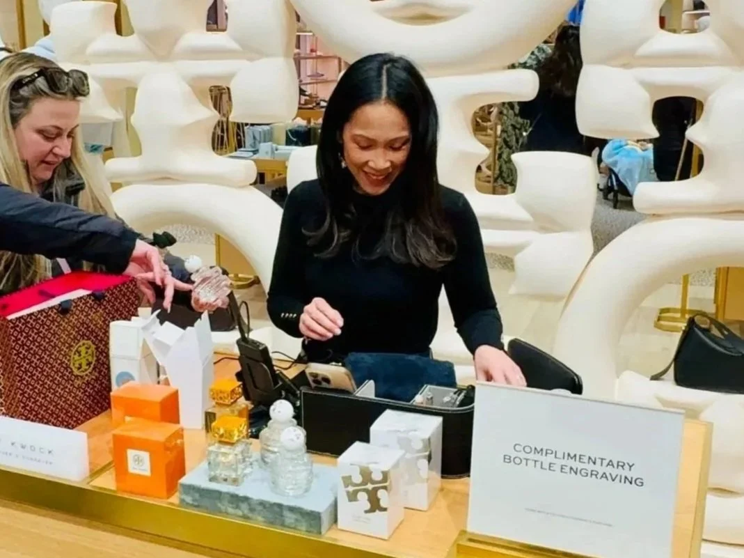 Woman smiling at a table in a retail store with perfume bottles and gift boxes, with a sign reading 'Complimentary Bottle Engraving'.