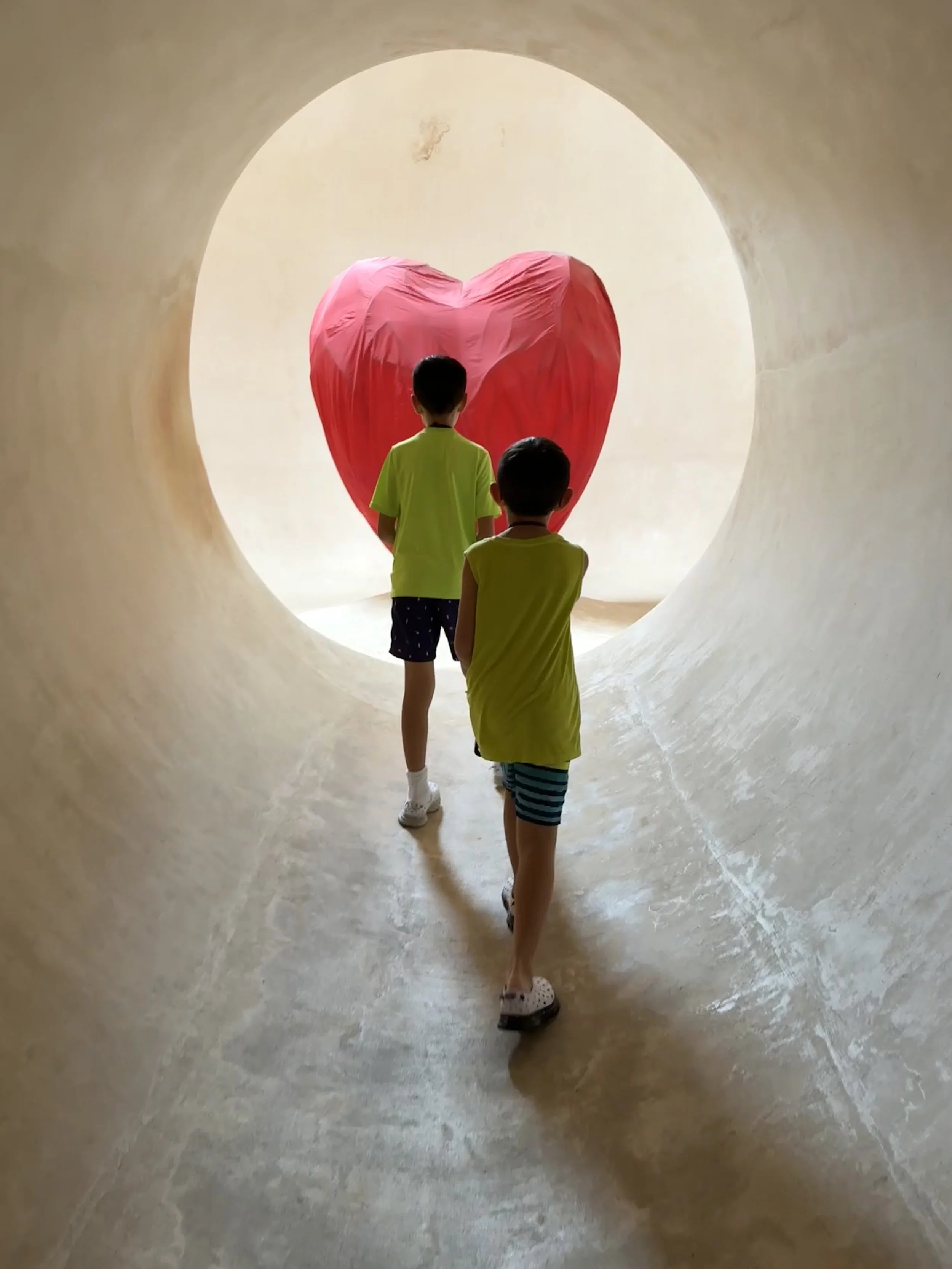 Two children walking through a circular tunnel towards a large red heart sculpture.