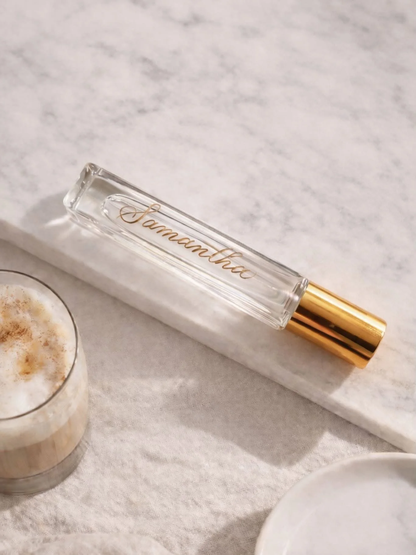 A clear glass perfume bottle with the name 'Saramasha' in gold script, placed on a white marble surface next to a candle and a partially visible white plate.