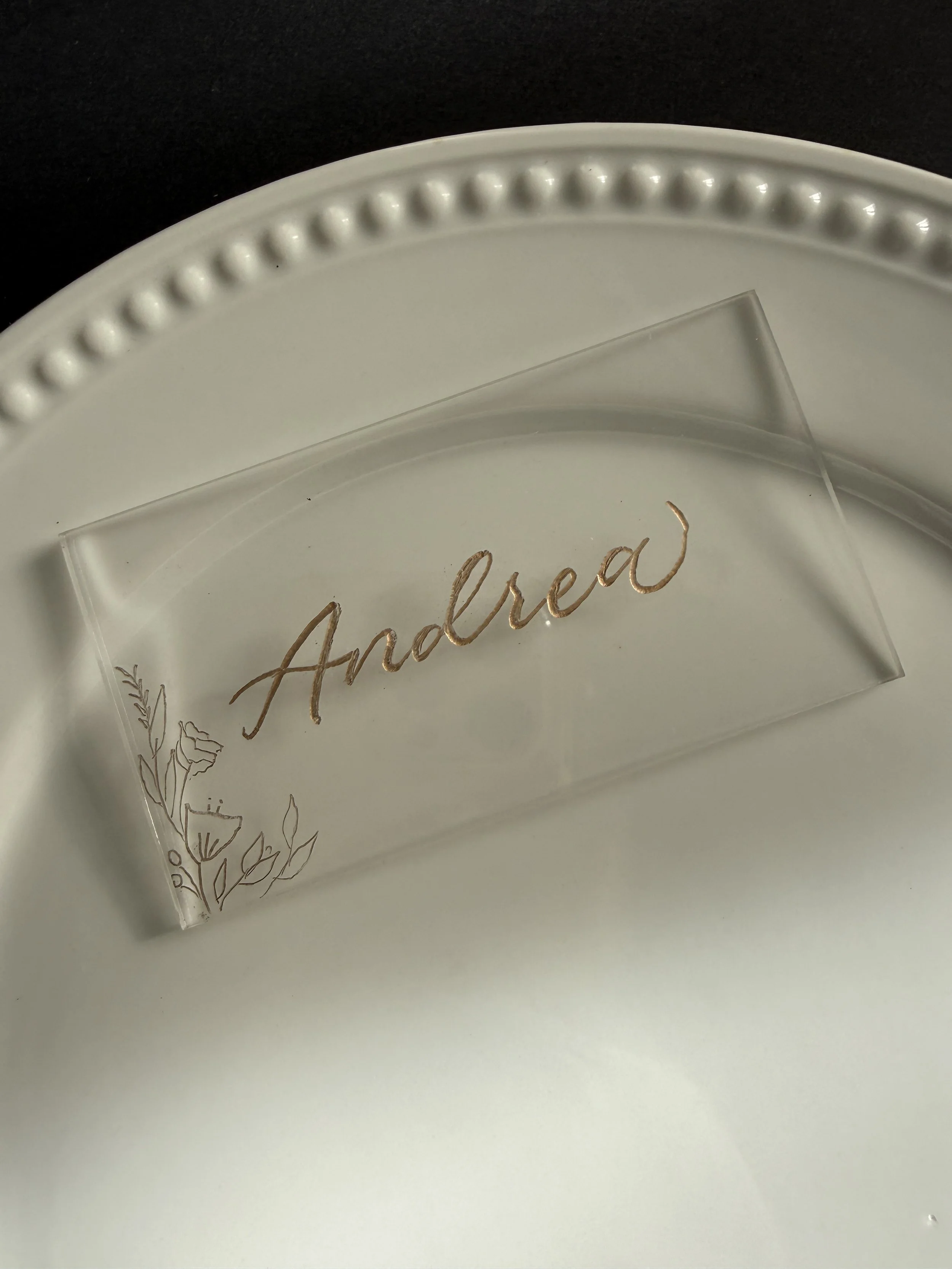 A clear acrylic nameplate with the name 'Andrea' written in gold cursive, placed on a white plate with a decorative beaded edge.