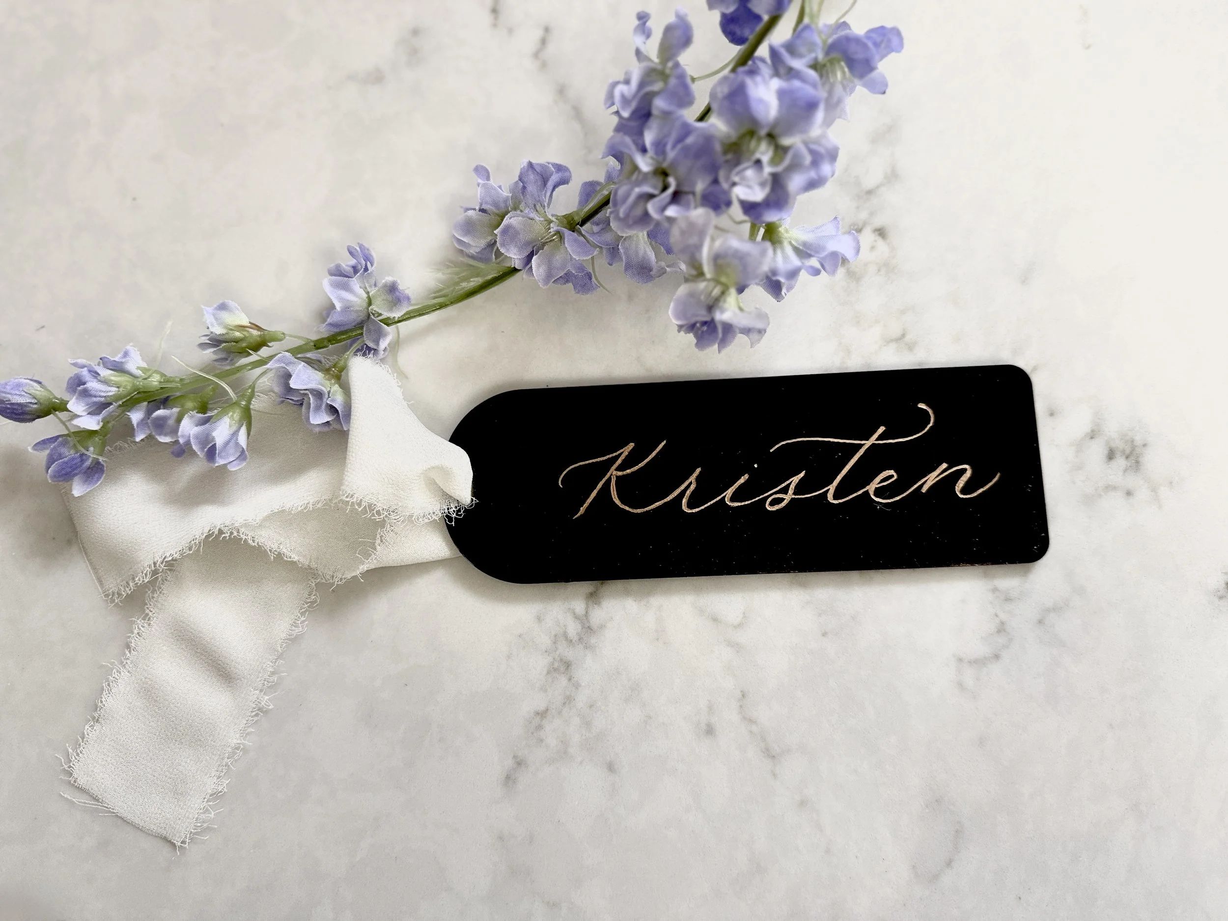 A sprig of light purple flowers with a white fabric ribbon and a black name tag with the name 'Kisten' written in cursive.