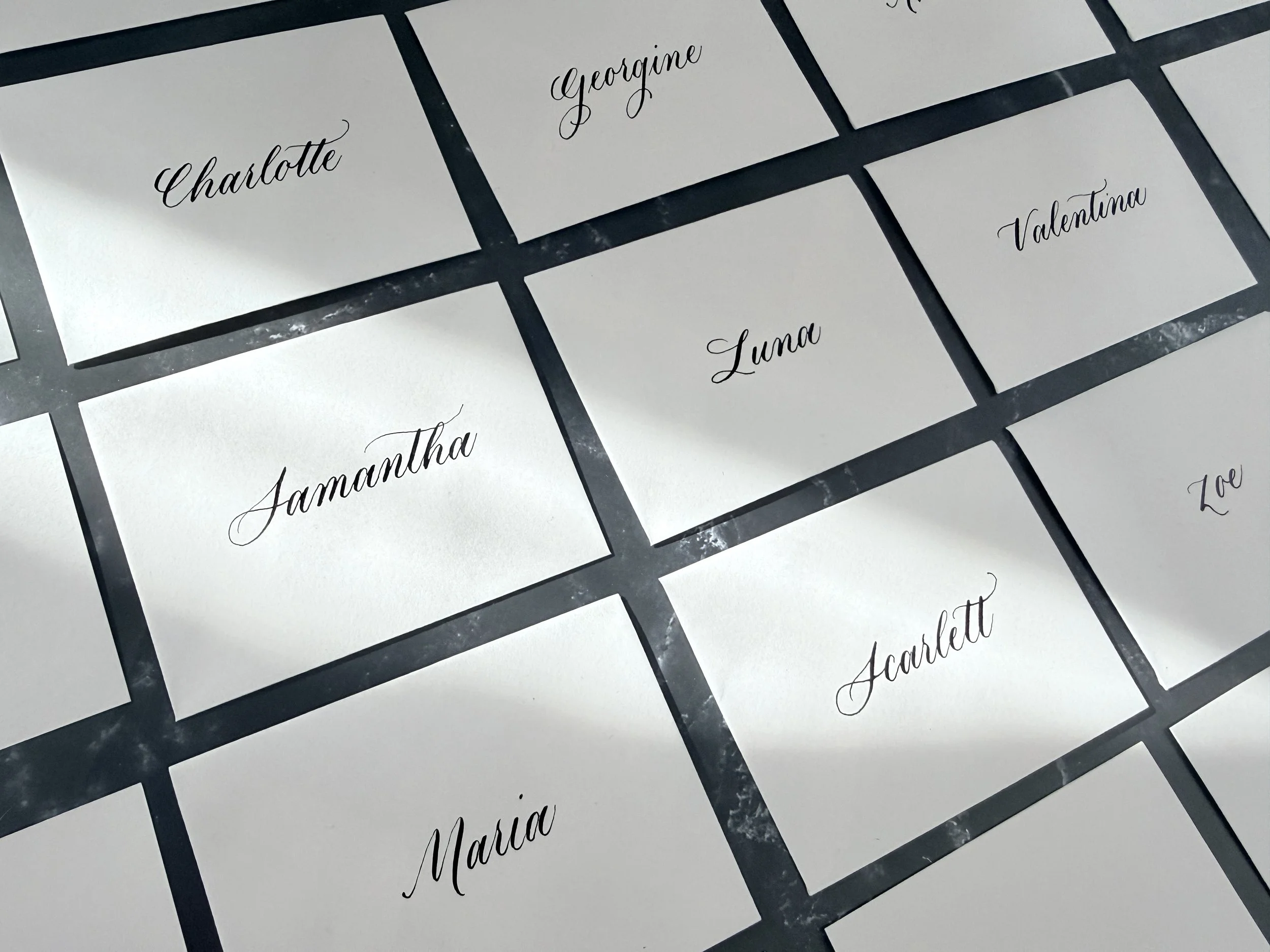 Name tags with cursive writing, including names Charlotte, Georgine, Valentina, Luna, Samantha, Scarlett, Maria, and Zoe, arranged on a dark surface.