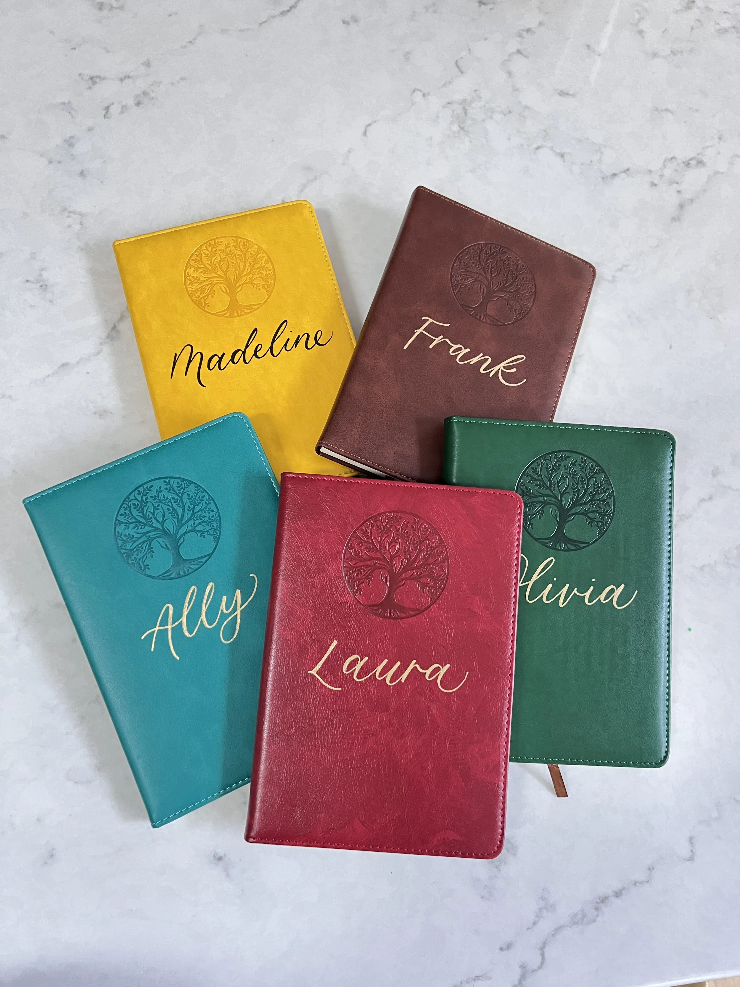Five colorful notebooks with a tree logo and names written on them, arranged on a white marble surface.