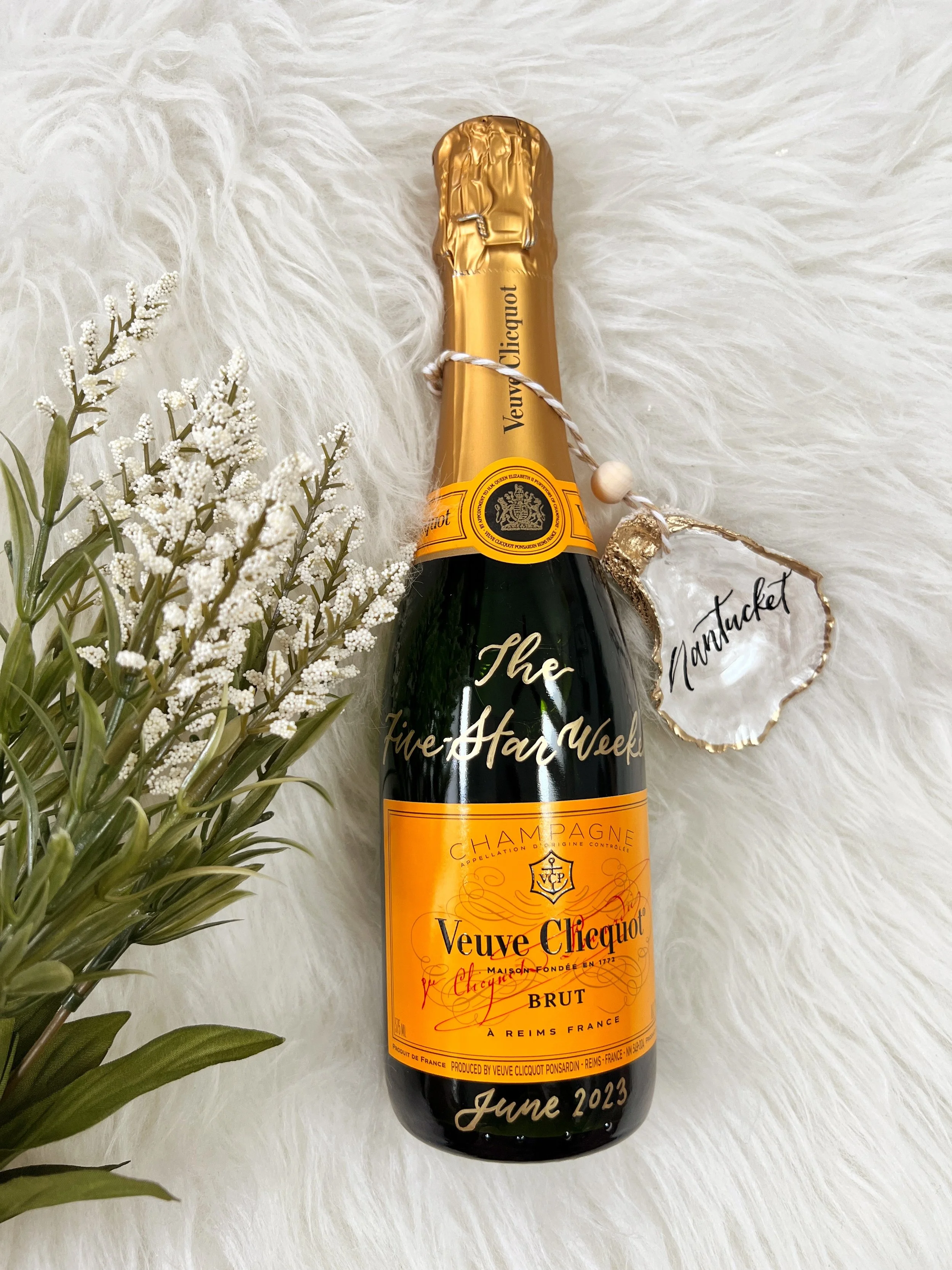 A bottle of Veuve Clicquot champagne with a gold foil top, placed on a white furry surface. Next to the bottle on the left, there is a small bouquet of white flowers. Hanging from the neck of the bottle is a string with a white bead, attached to a wh