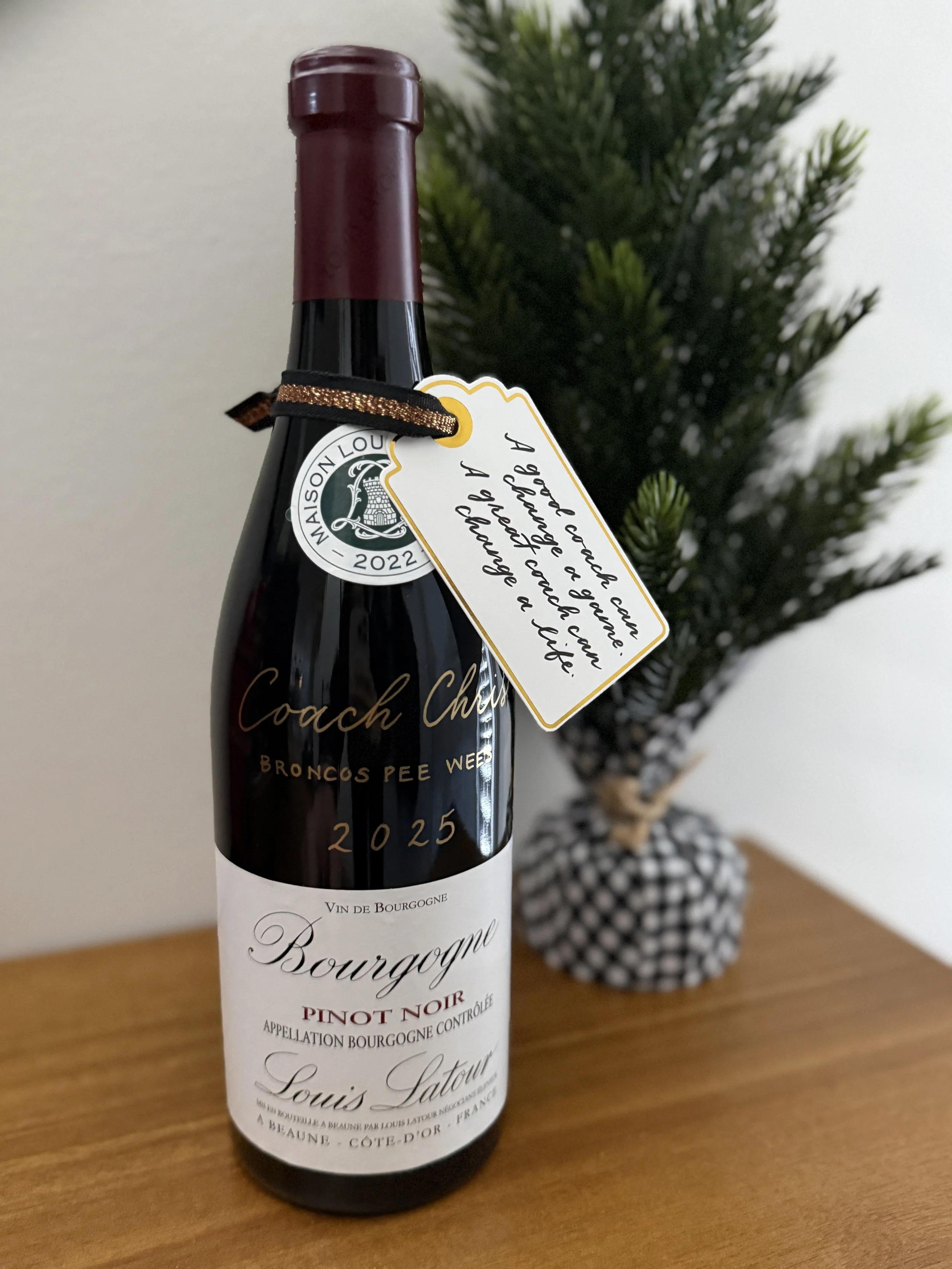 A bottle of Bourgogne Pinot Noir wine with a tag that says, "A great wine, a friend, a glass, and a smile are always good for me." Next to the bottle is a small potted pine tree on a wooden surface.