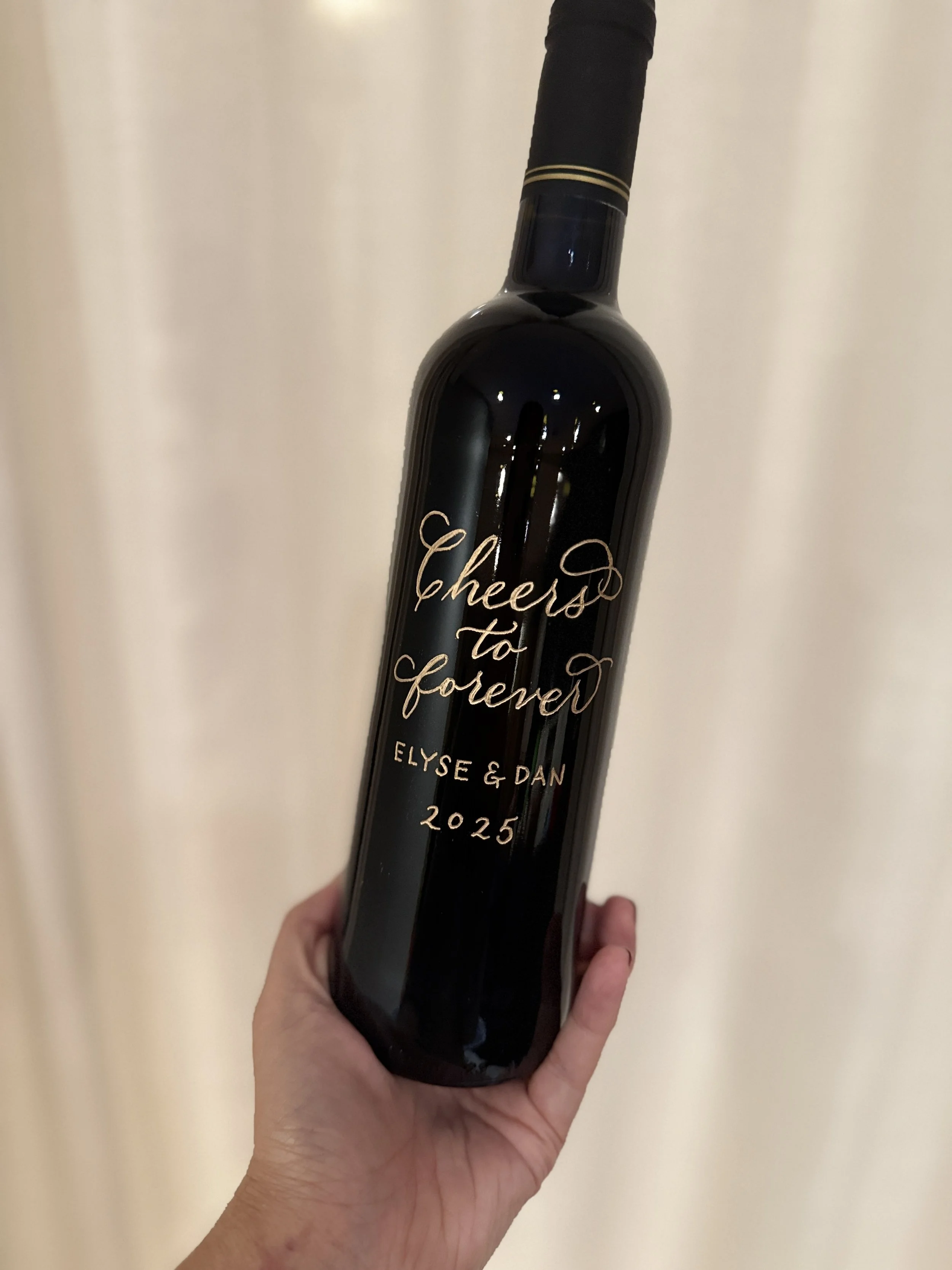 A hand holding a black wine bottle with a personalized message in gold lettering that says, 'Cheers to forever,' with the names Elyse & Dan and the year 2025.