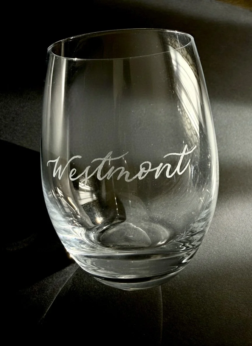 A clear glass with the word 'Westmont' etched on it, placed on a dark surface with a dark background.