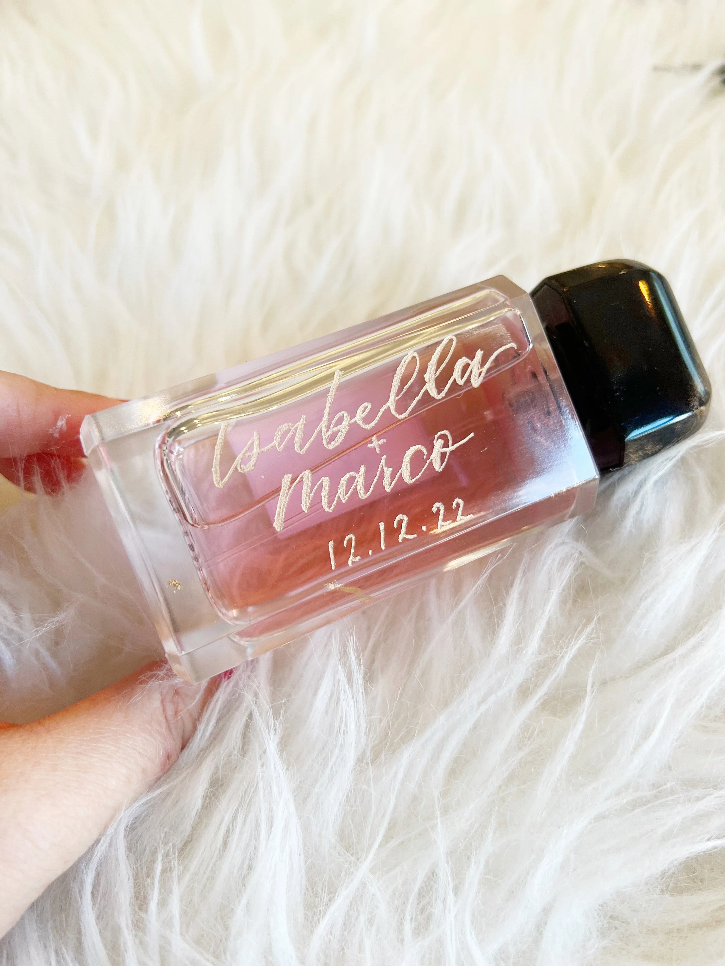 Clear glass bottle with pink liquid inside, engraved with names Isabella and Marco, and date 12.12.22, resting on a white, fluffy surface.