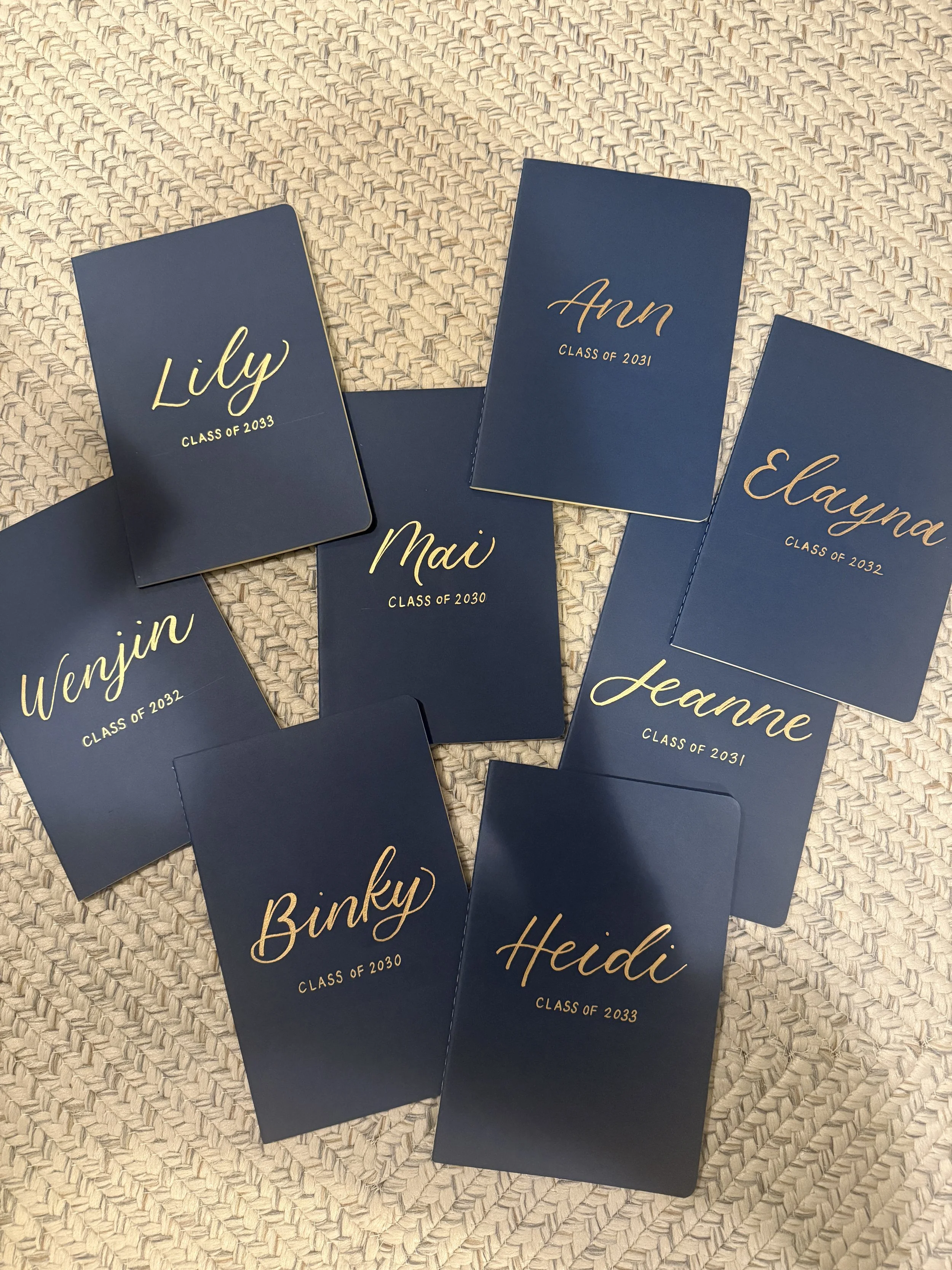 A collection of navy blue graduation cards with gold cursive names and class years, laid out on a woven beige mat.