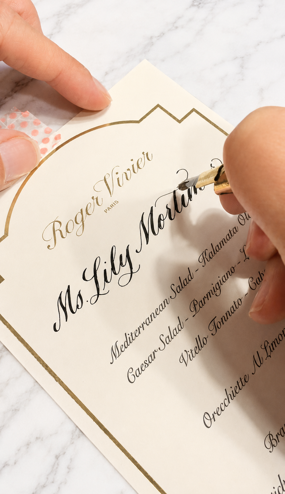 Person writing calligraphy on a wedding menu card from Roger Vivier Paris, with a marble surface in the background.
