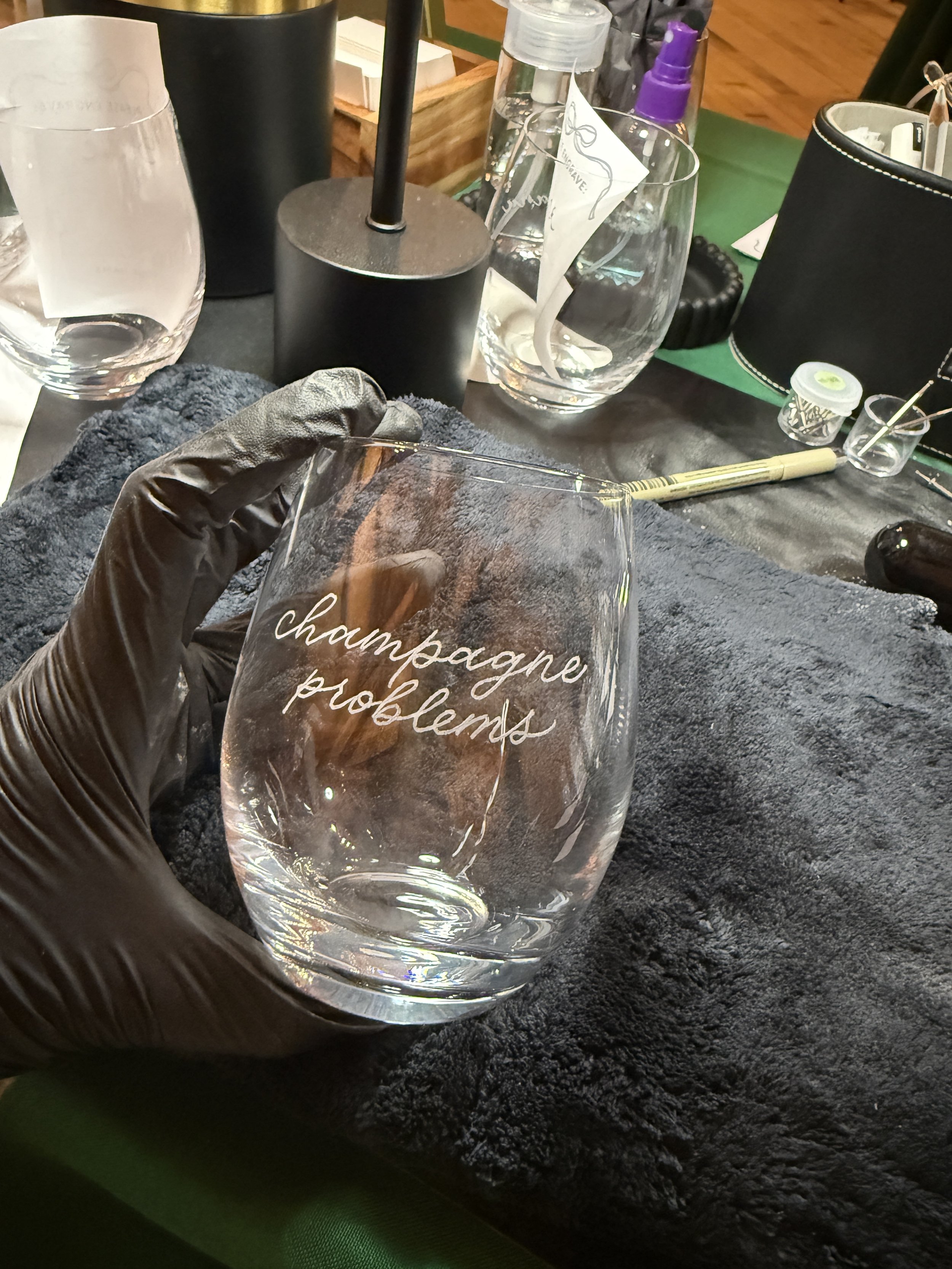 A glass with the words 'champagne problems' etched on it being held by a hand wearing a black glove. The background shows various bottles, tools, and supplies on a work surface.