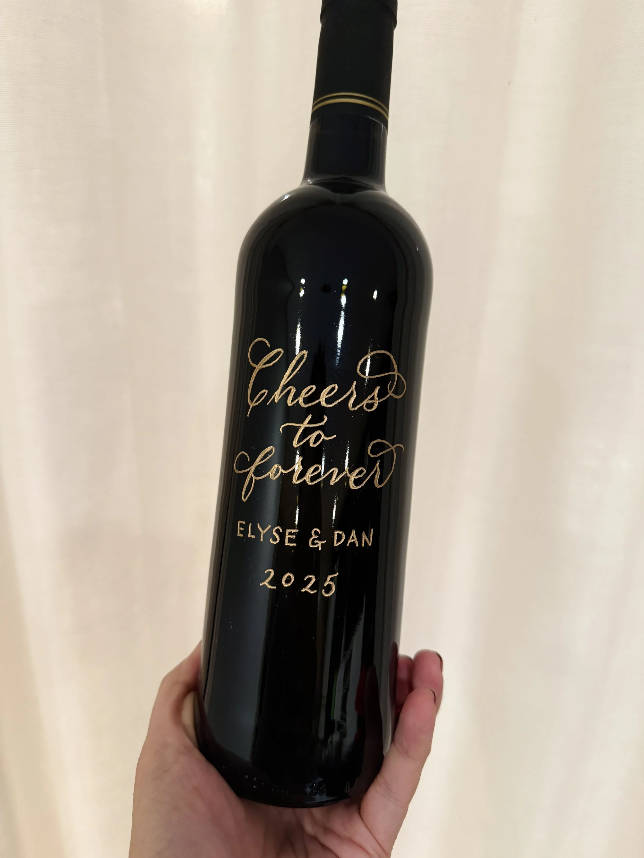 A black wine bottle with gold writing that says "Cheers to forever" and includes the names Elyse & Dan and the year 2025.