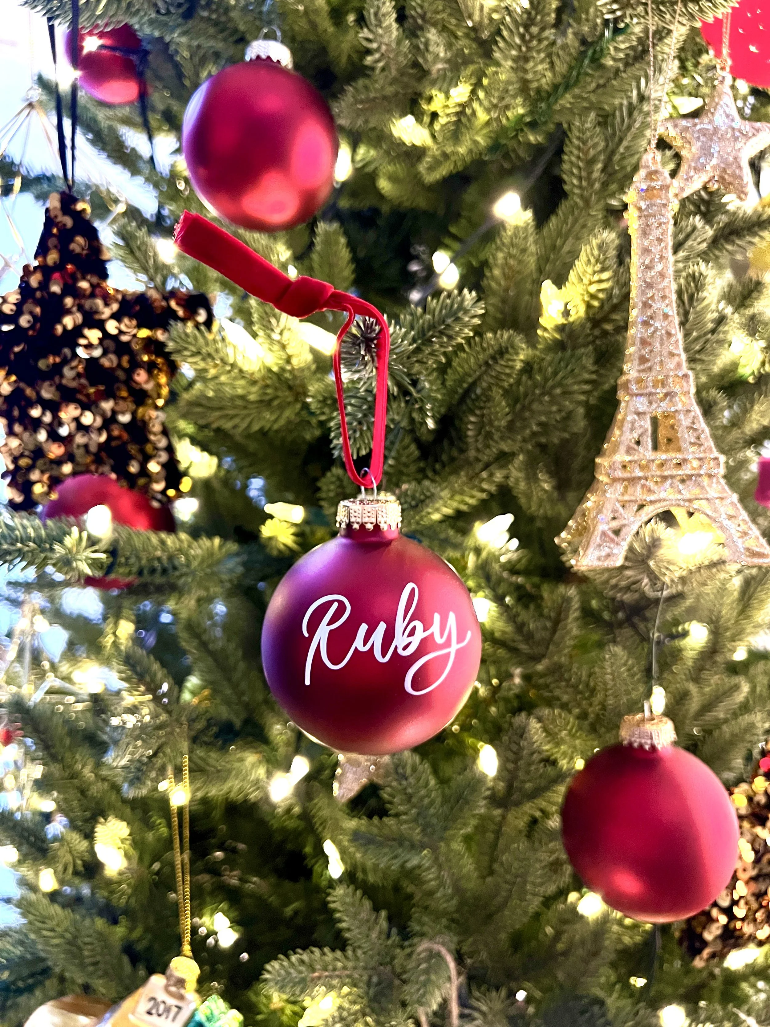 Close-up of a Christmas tree decorated with red ornaments, including a spherical ornament with the name 'Ruby' written on it, a gold Eiffel Tower ornament, and other festive decorations, illuminated with warm white string lights.