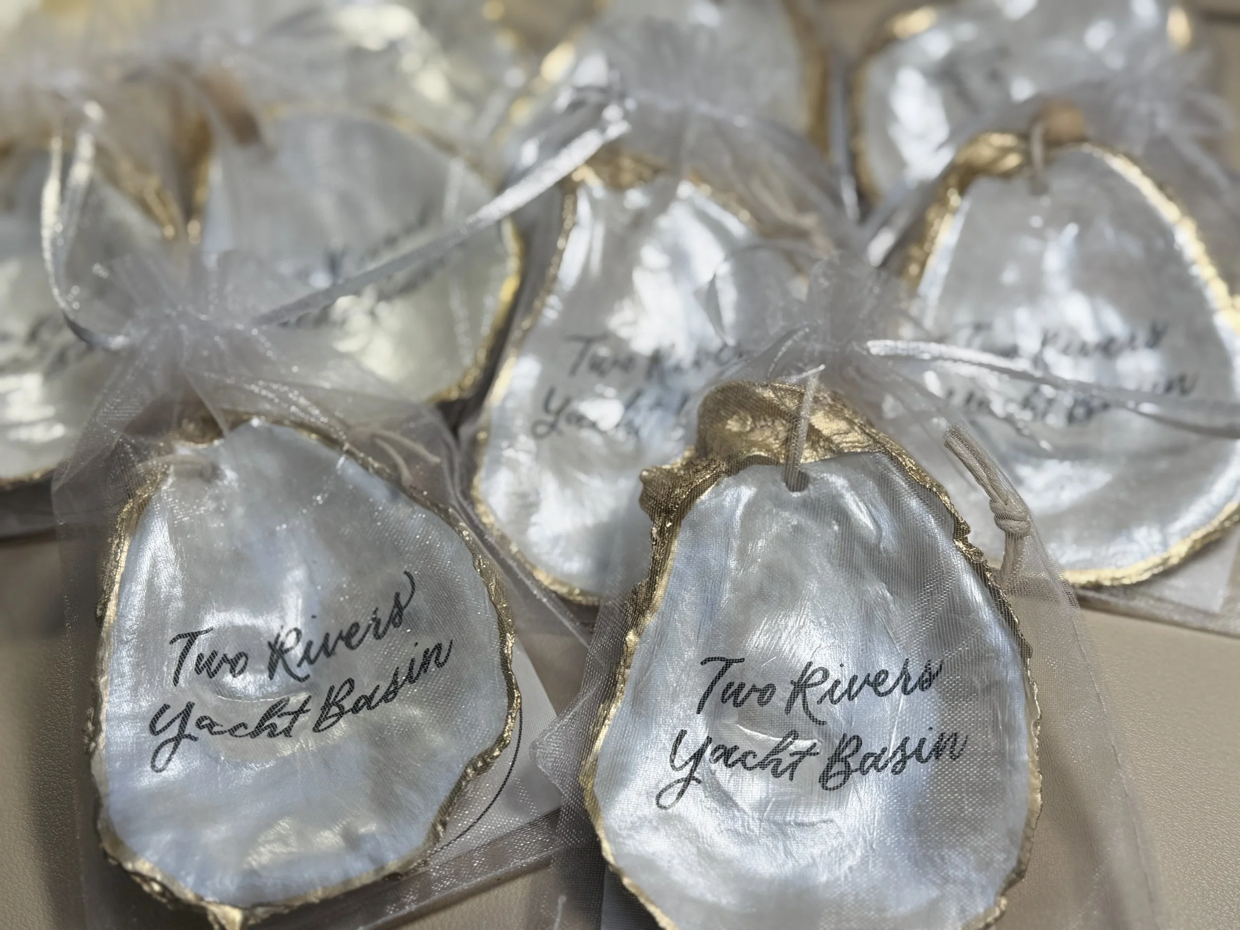 Decorative oval-shaped items with white satin centers, gold trim, and black cursive text reading 'Two Rivers Yacht Bash', wrapped in sheer white material with gold edges.