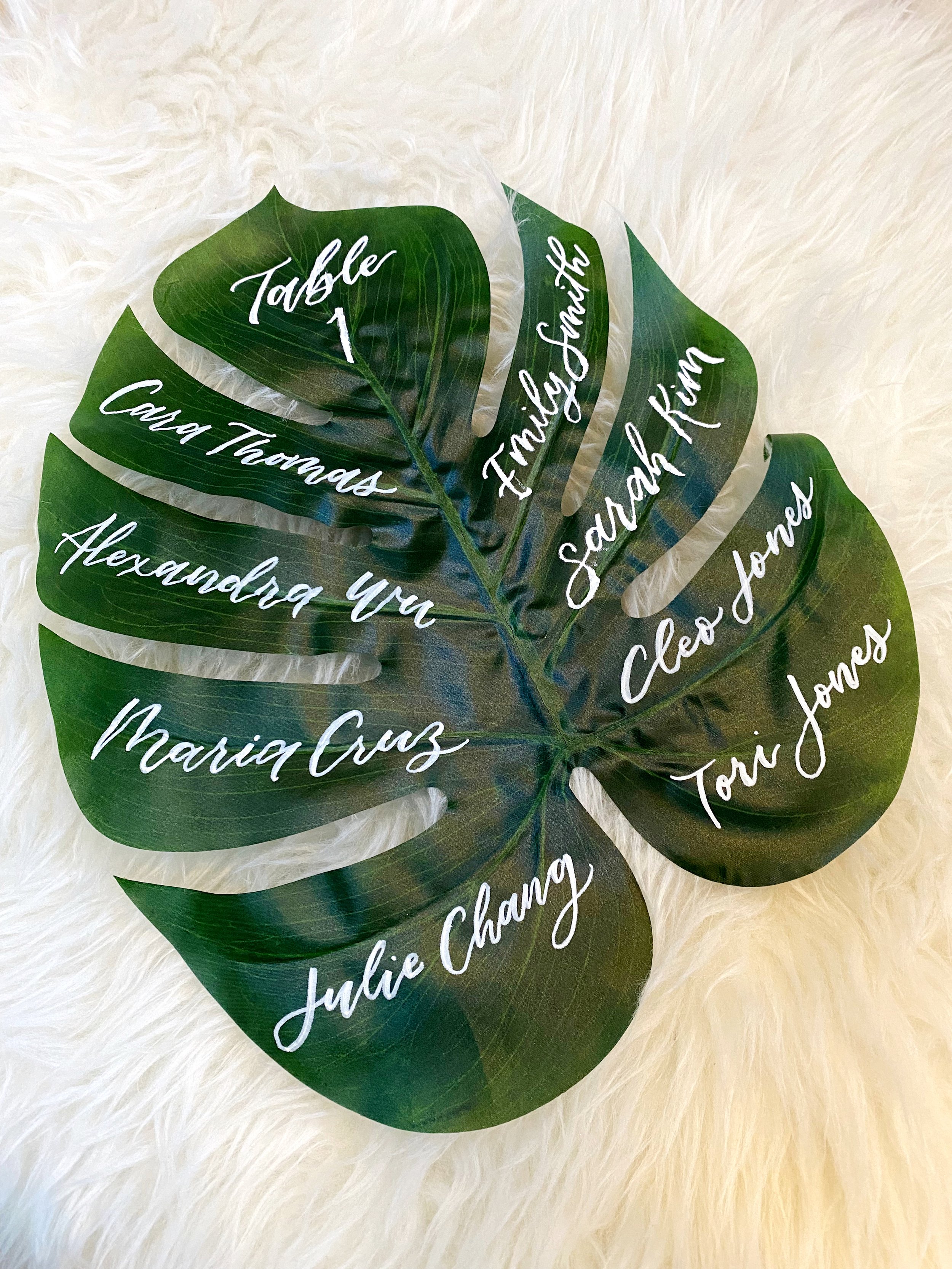 Decorative tabletop name card with a large green tropical leaf and white handwritten names and table number.