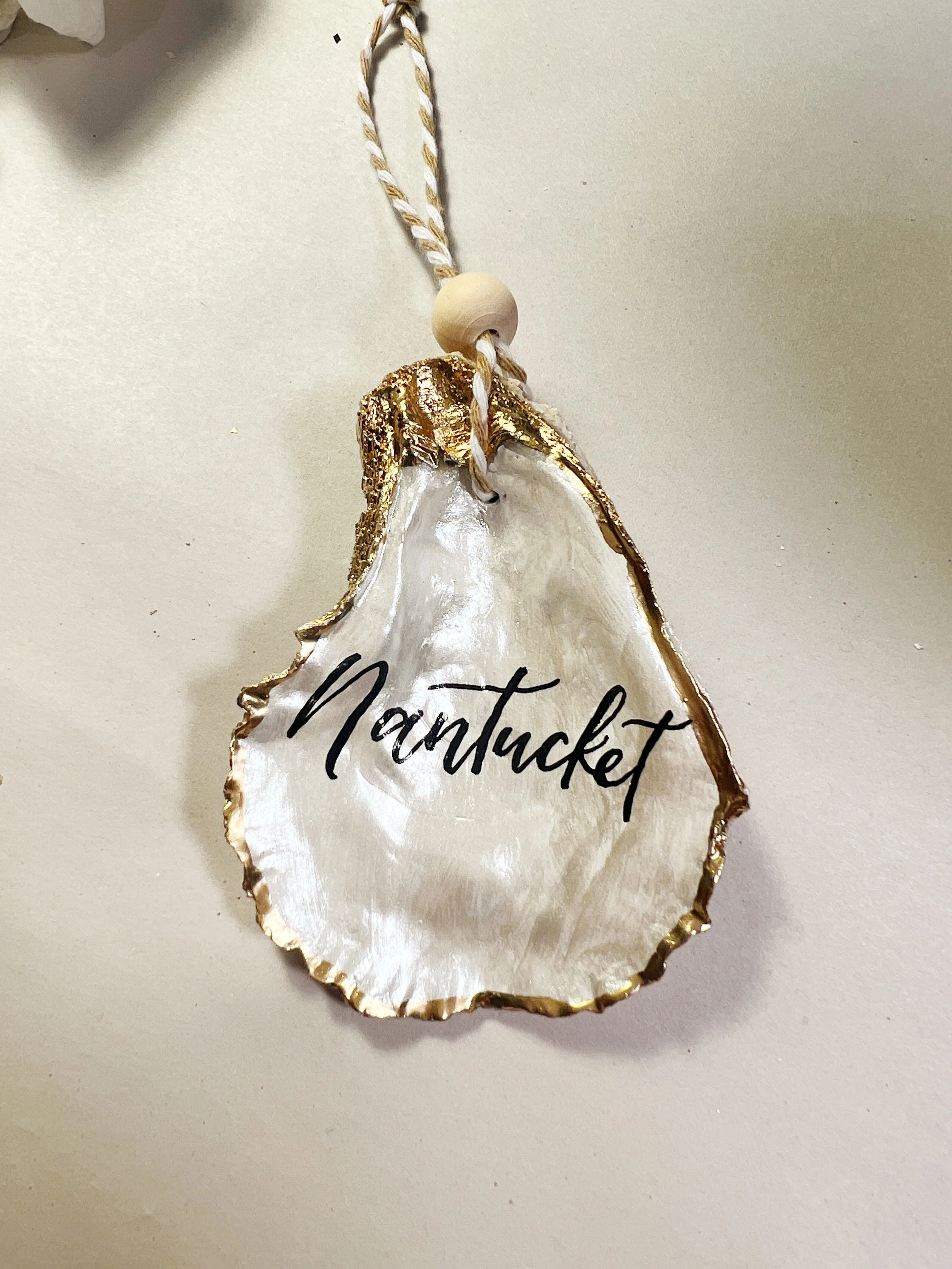 Decorative shell ornament with the word 'Nantucket' written on it, hanging from a white and gold string.