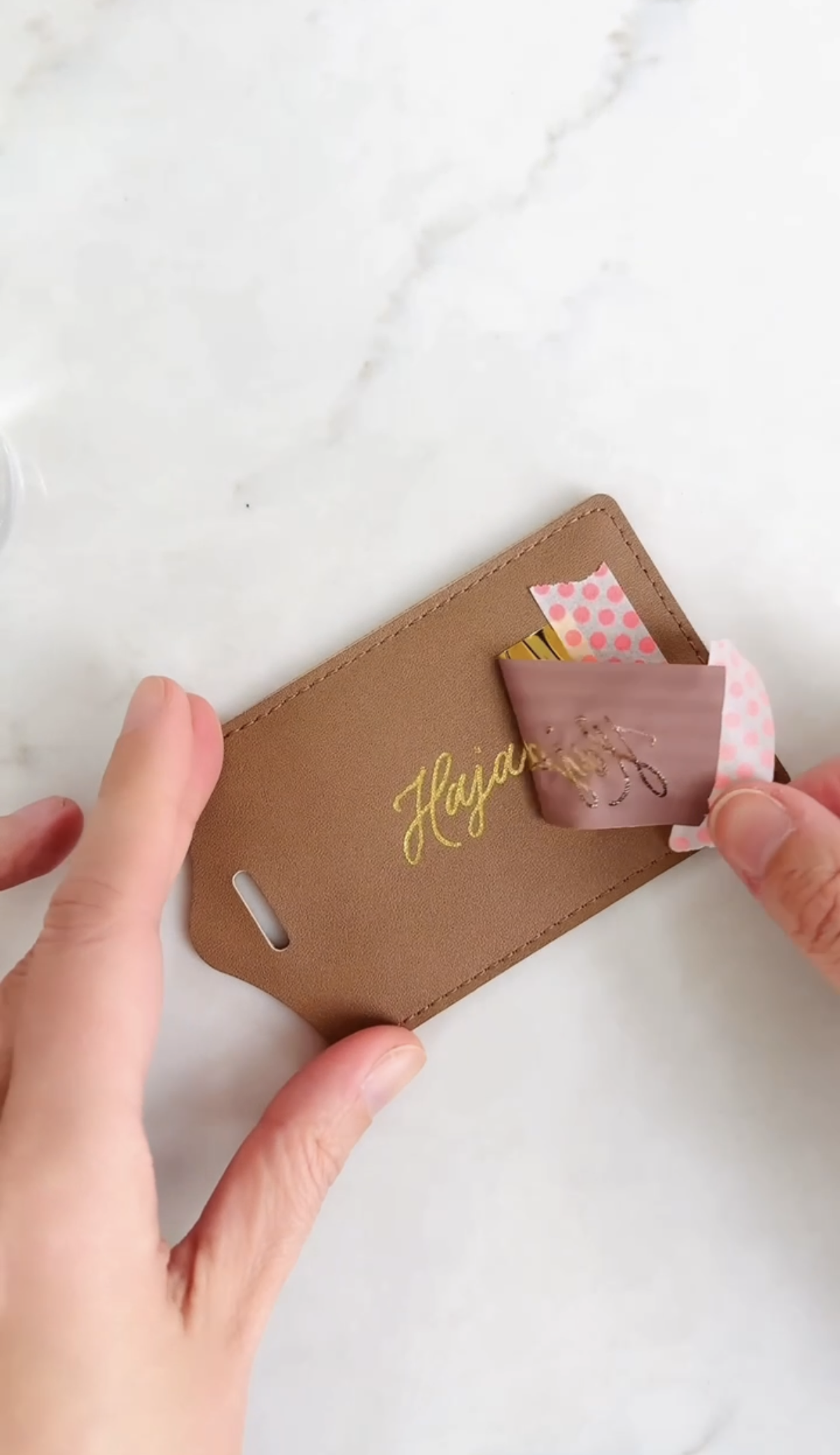 Person removing a decorative pink and gold sticker from a tan cardholder with the word 'Hayden' embroidered in gold.