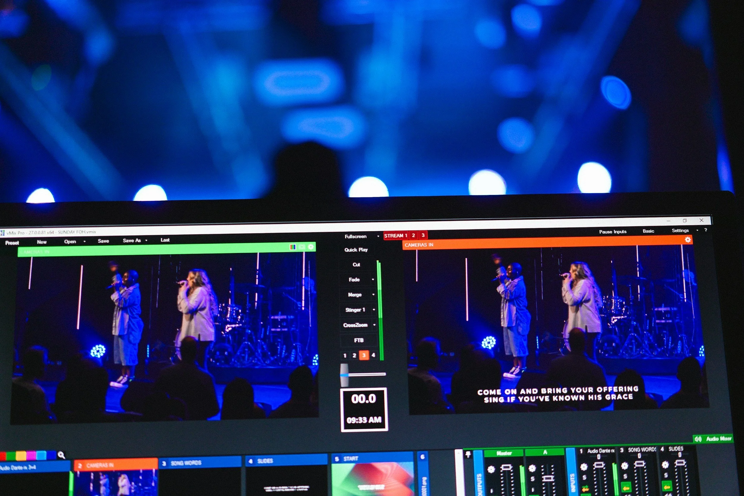A computer screen displaying a live video feed of a singer and a backup vocalist performing on stage, with stage lights and musical instruments in the background, and lyrics at the bottom reading 'Come on and bring your offering, sing if you've known His grace'.