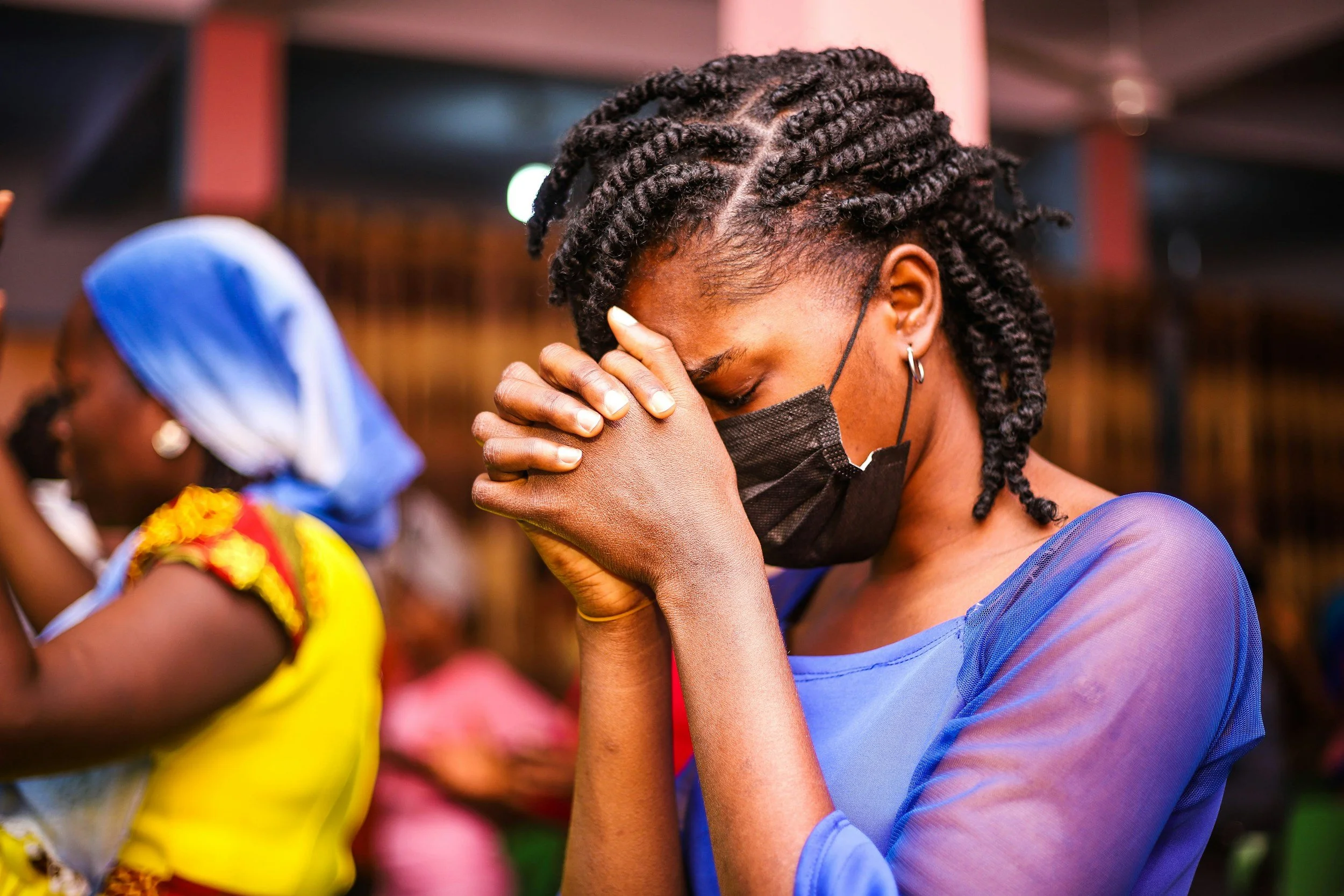 A woman wearing a black face mask, a blue sheer top, and earrings prays with her hands clasped and head bowed. In the background, another woman wearing traditional clothing and a headwrap also prays with her hands raised.