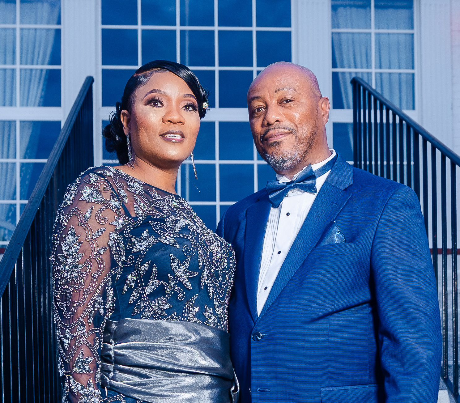 A well-dressed couple in formal evening attire standing on a staircase outside a modern building with large glass windows.