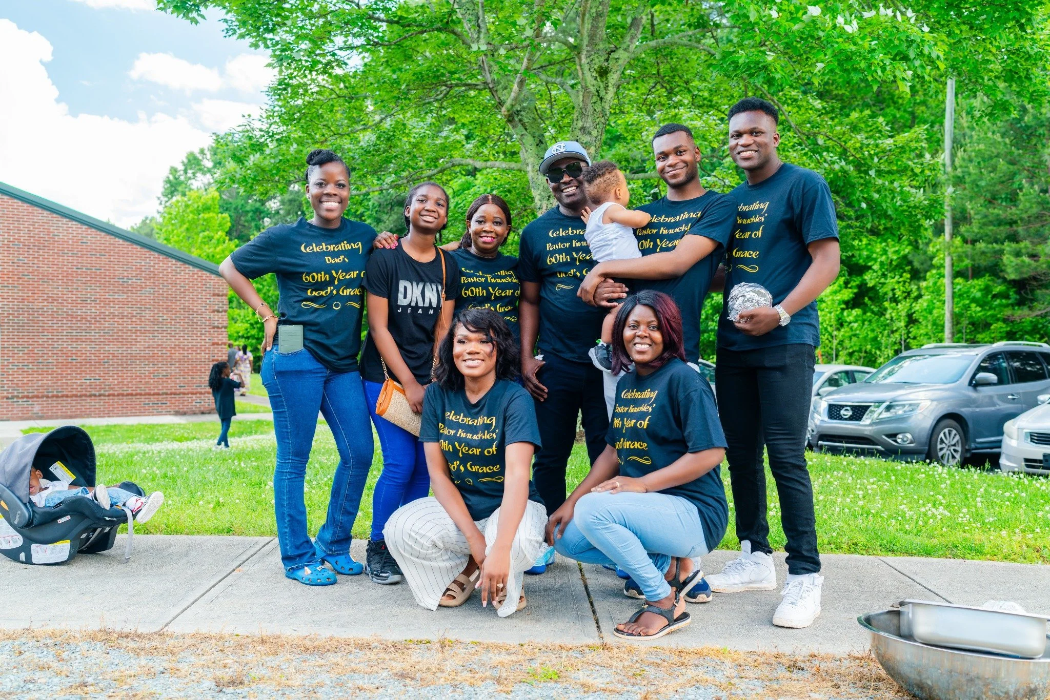 Family gathering outdoors celebrating a 60th birthday, with most wearing matching black t-shirts that read 'Celebrating Pastor Knuckles 60th Year of God's Grace.' They are smiling, posing on a sidewalk with green trees and parked cars in the background.