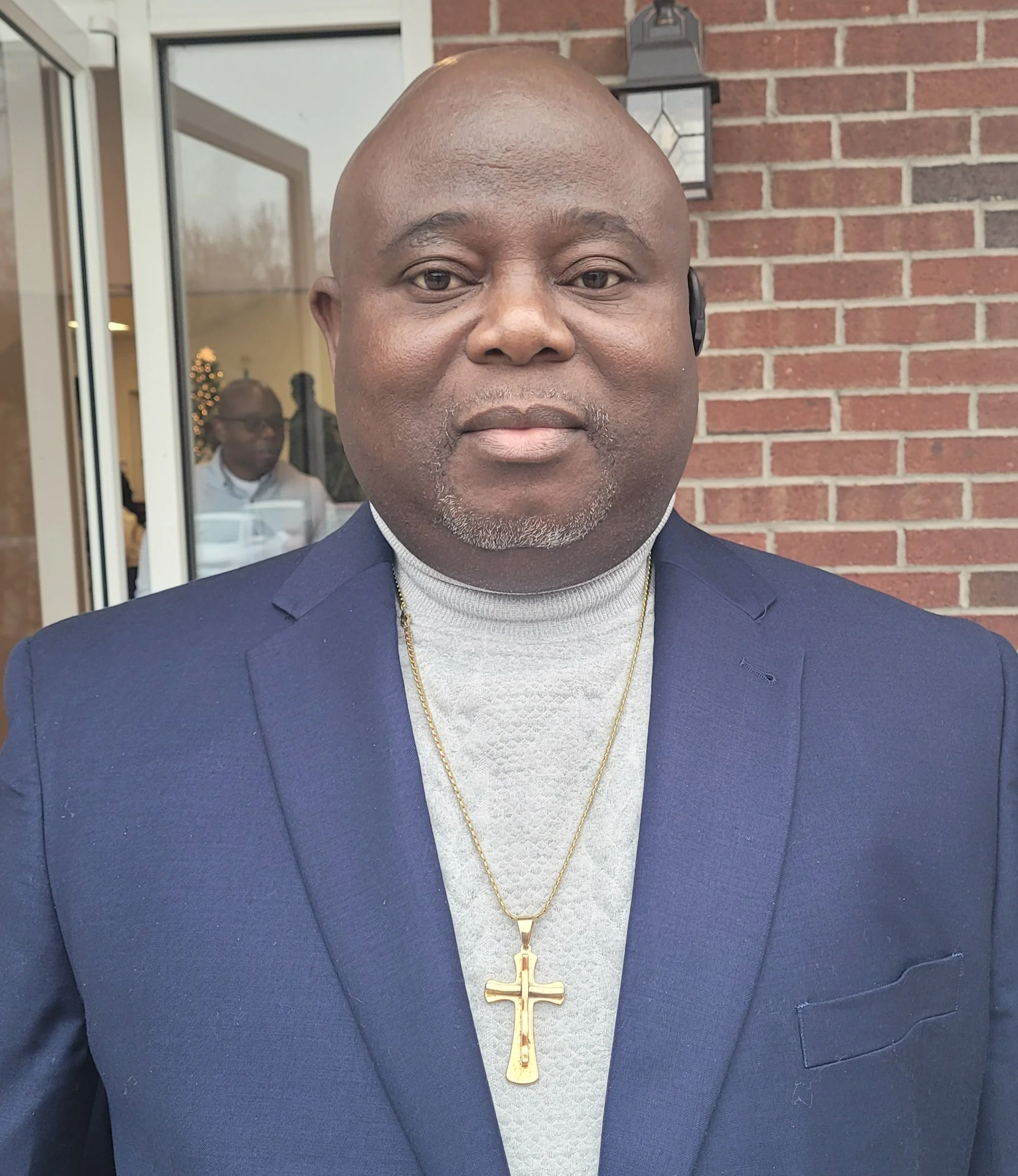 A man wearing a dark blue suit with a white turtleneck and a gold cross necklace standing outside in front of a brick wall, with a woman visible through the glass door behind him.