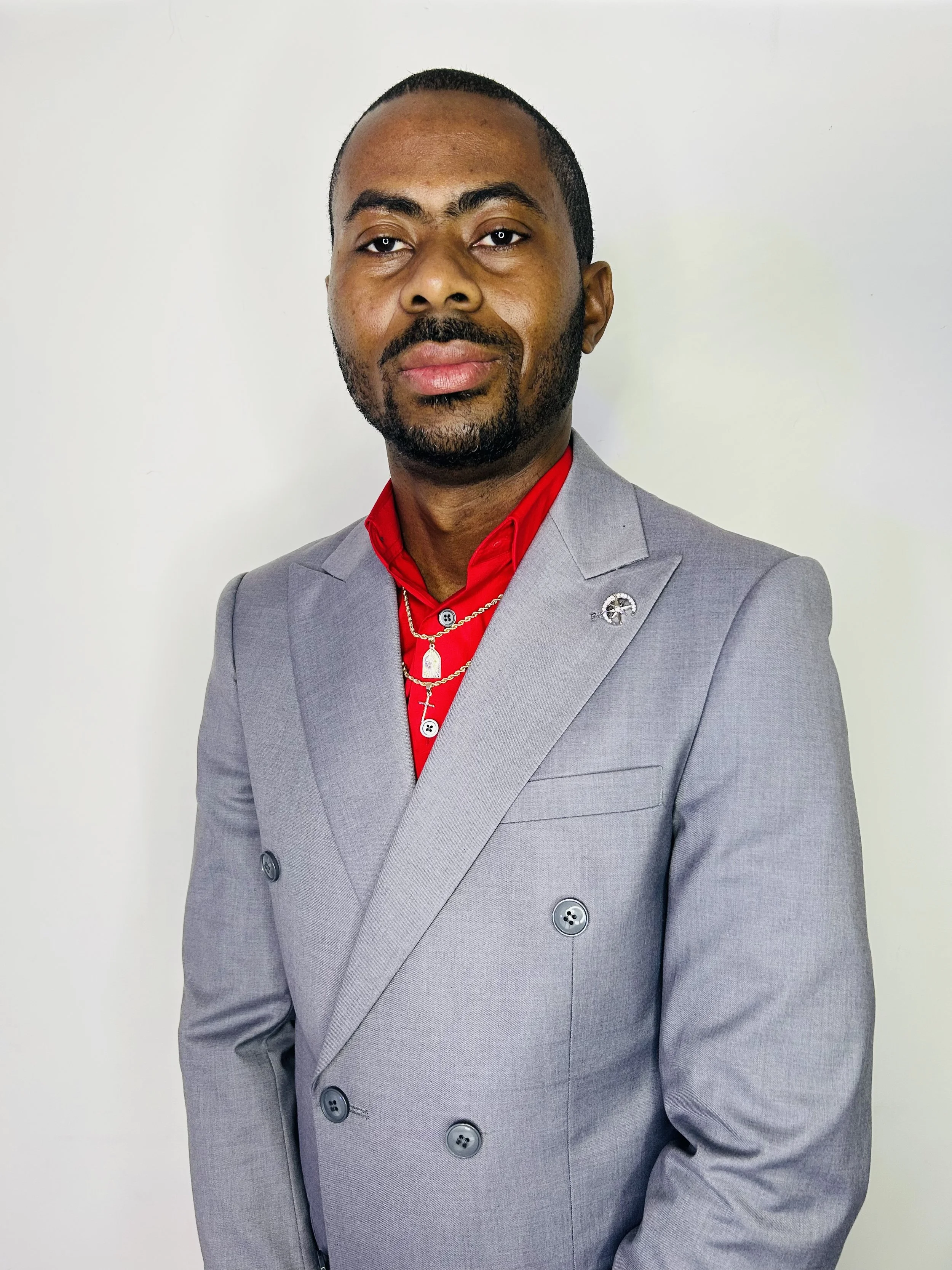 A man with a beard and mustache wearing a gray double-breasted suit jacket, red shirt, and layered gold necklaces, standing against a plain, light-colored background.
