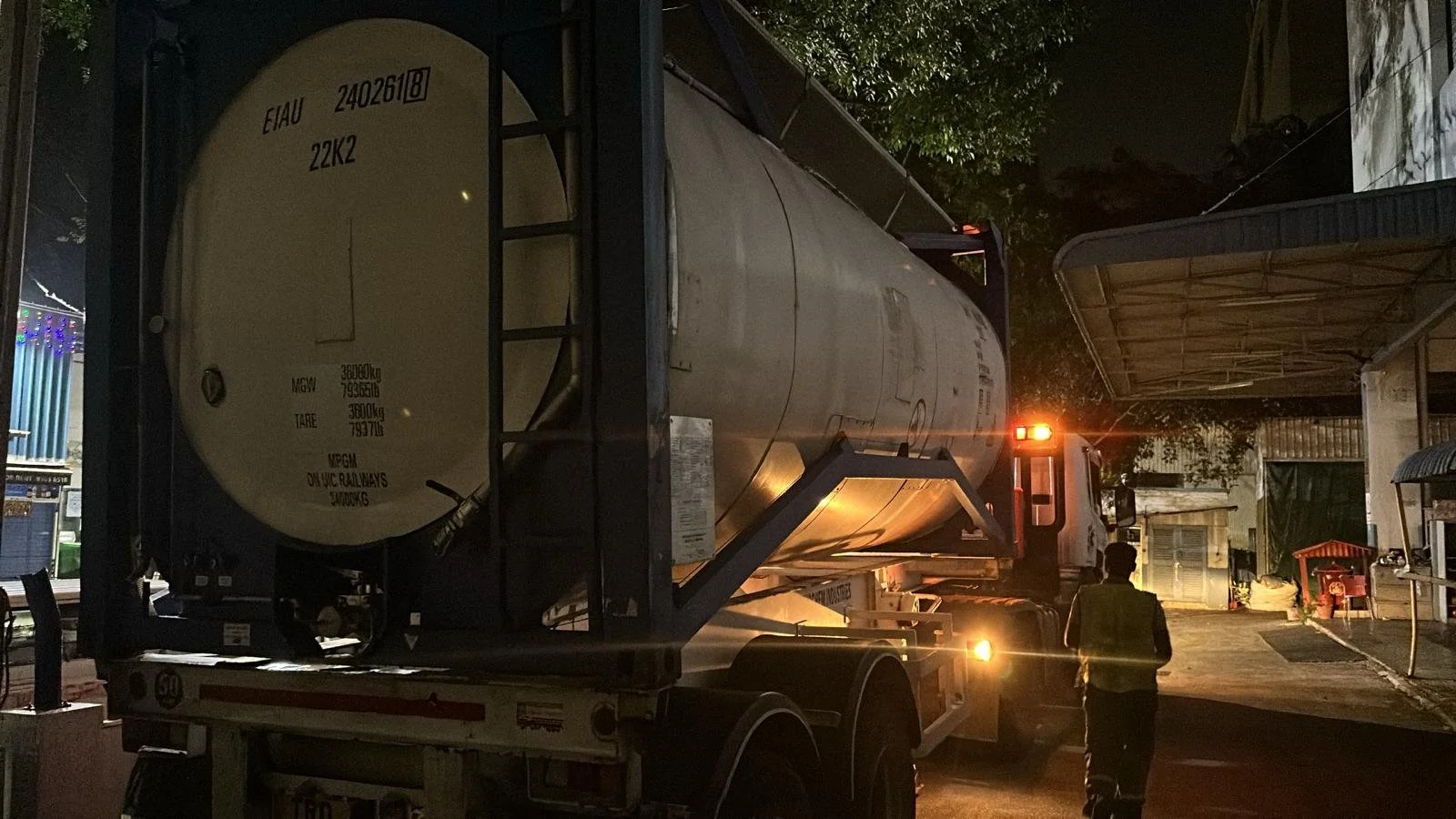 A night scene showing a large tanker truck on a street with a person in a reflective vest walking nearby. The truck has a cylindrical tank with various markings, and there are street lights and trees in the background.