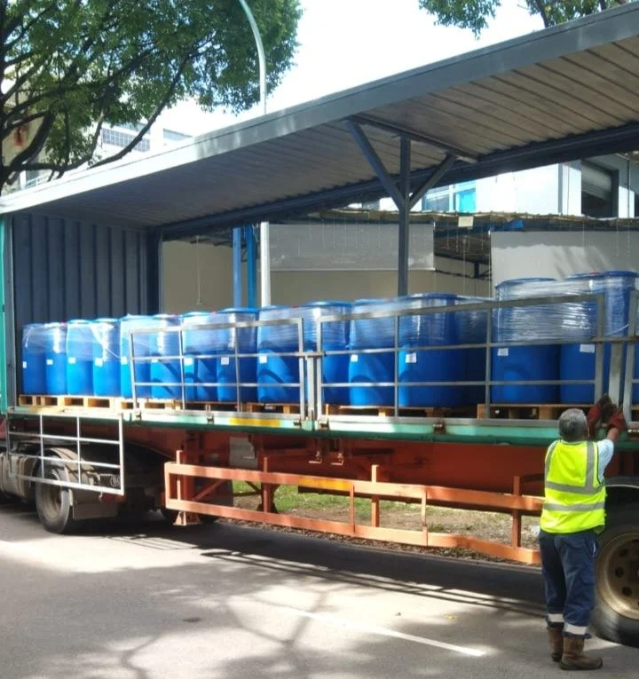 A semi-truck with an open trailer carrying large blue plastic barrels, with a worker in a yellow safety vest standing beside the truck.