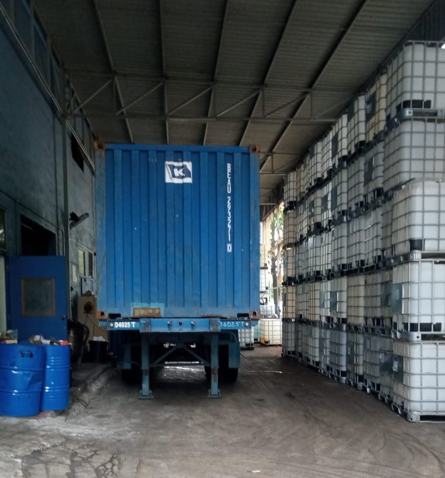 A blue shipping container on a flatbed truck inside a warehouse, with stacked white intermediate bulk containers on the right and blue drums on the left.
