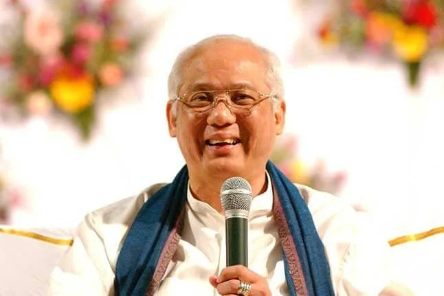 A smiling elderly man with glasses holds a microphone and wears a white shirt and a blue scarf, with blurred colorful flowers in the background.