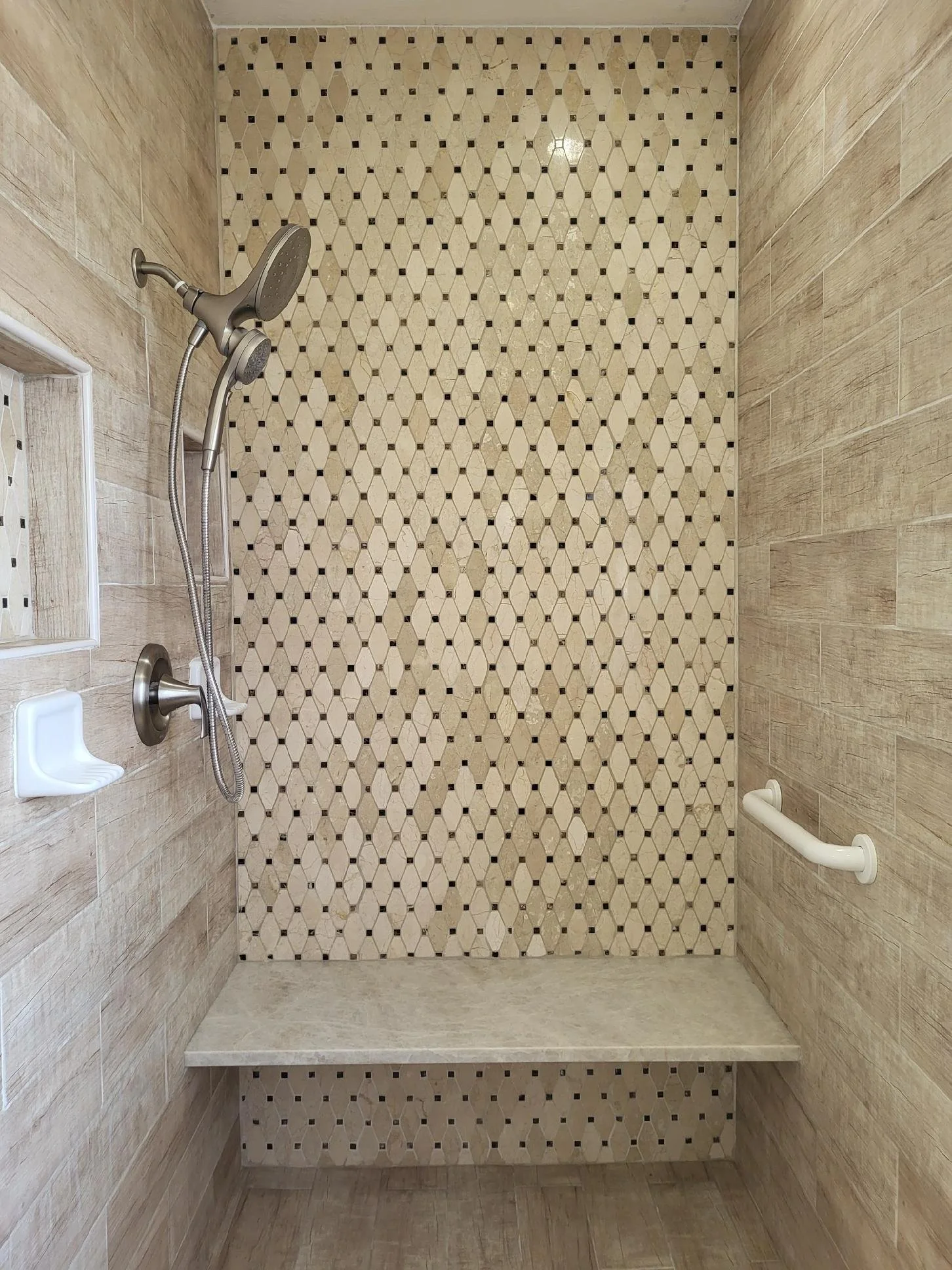 Shower with a beige and black geometric tile accent wall, a bench, a soap dish, a handheld showerhead, and a white grab bar.