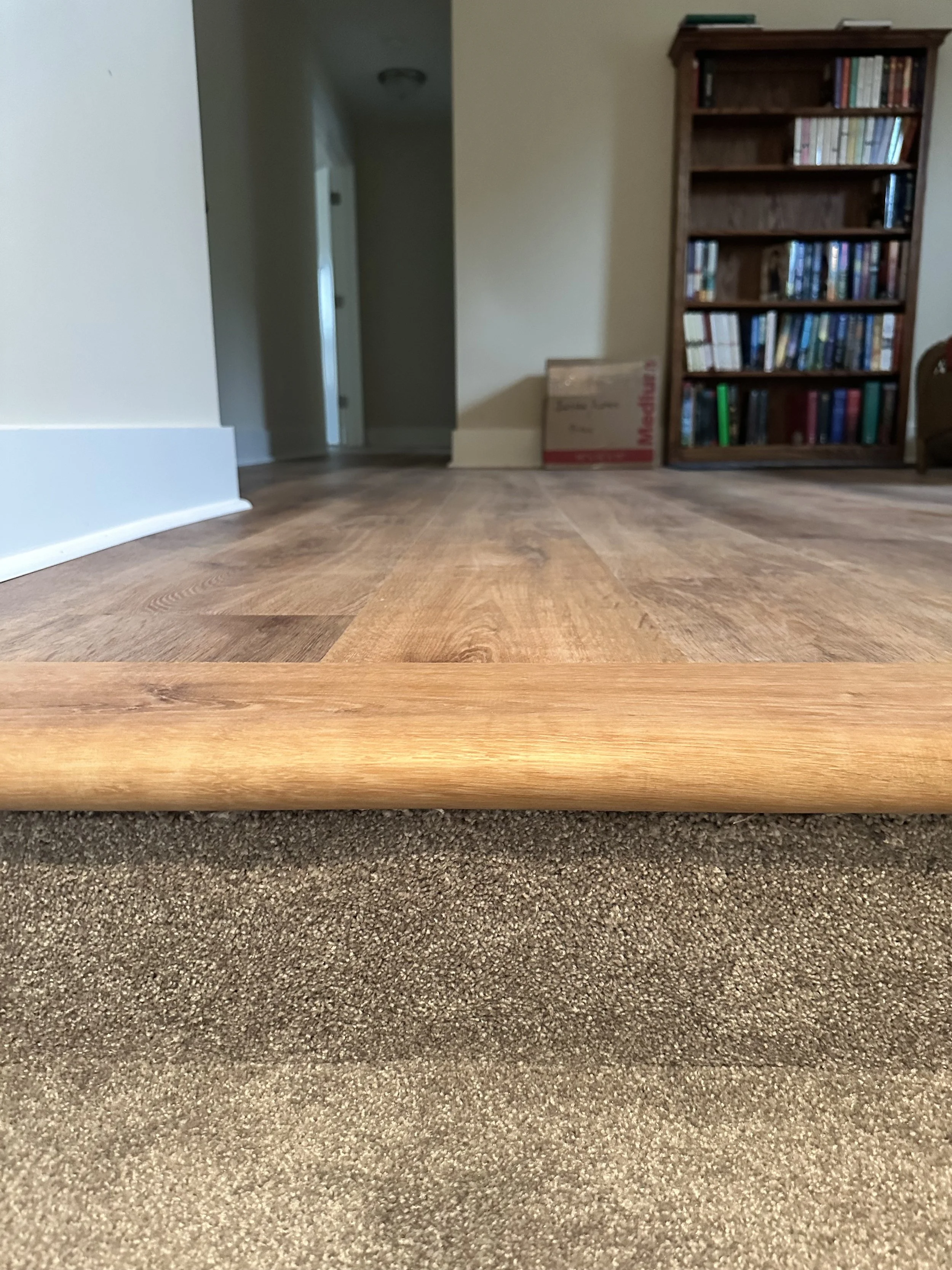 View of a living room from a low angle showing a transition between beige carpet and hardwood floor, with a wooden bookshelf and living room doorway in the background.