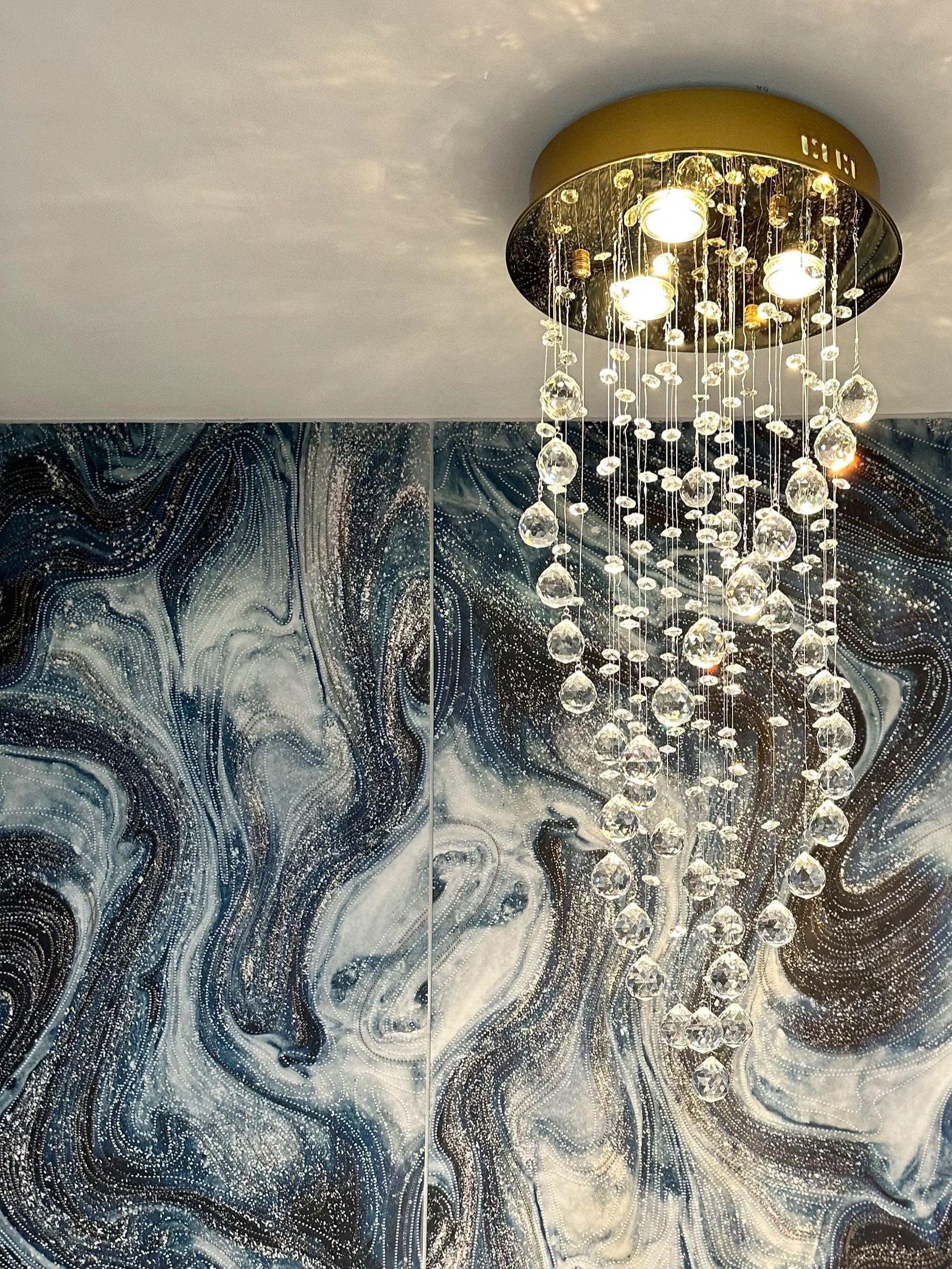 Elegant ceiling chandelier with multiple hanging crystal-like ornaments illuminating a modern, marbled wall with swirling black, white, and grey patterns.