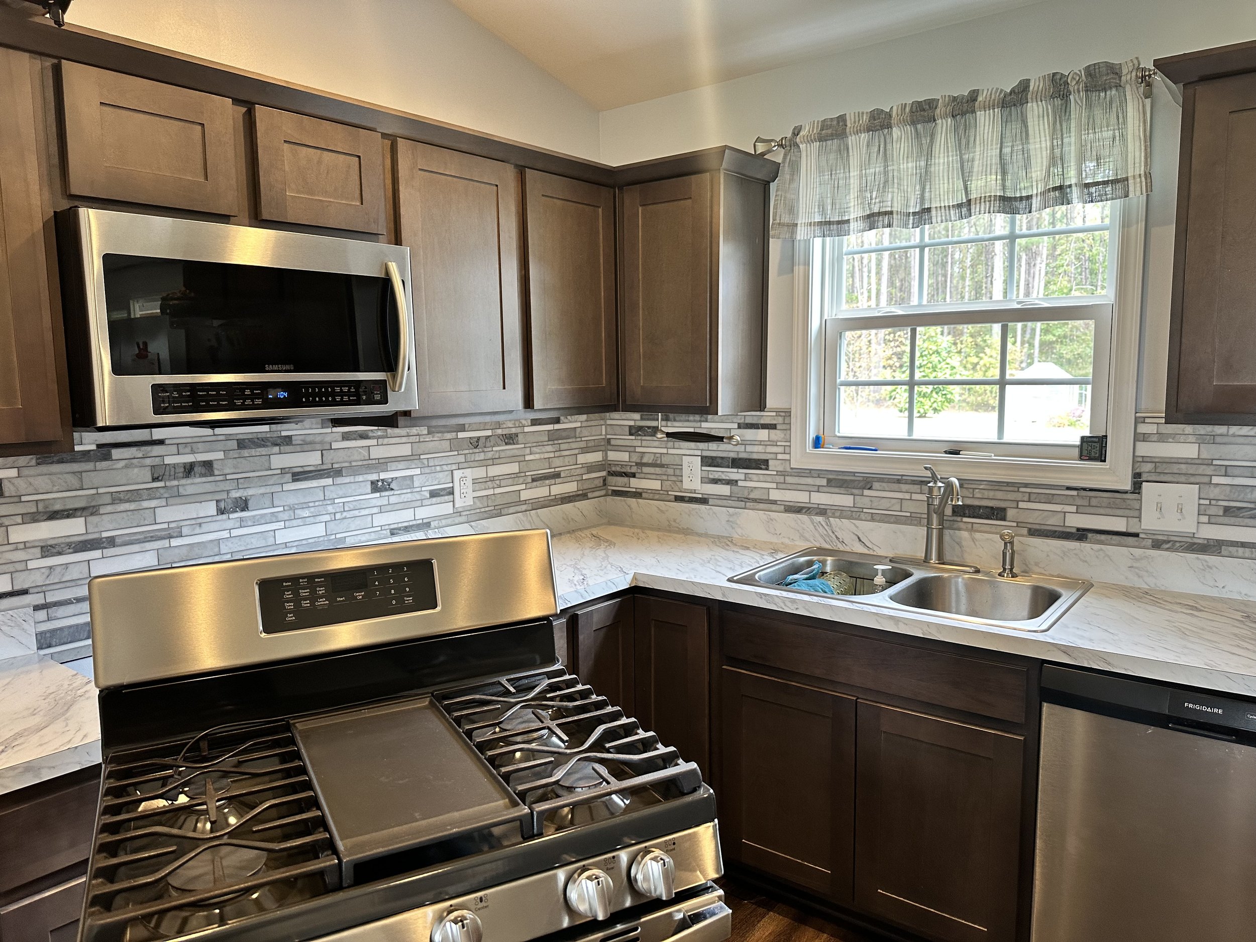 Kitchen with brown cabinets, marble countertop, mosaic tile backsplash, stainless steel microwave and stove, sink with window above, and dishwasher.