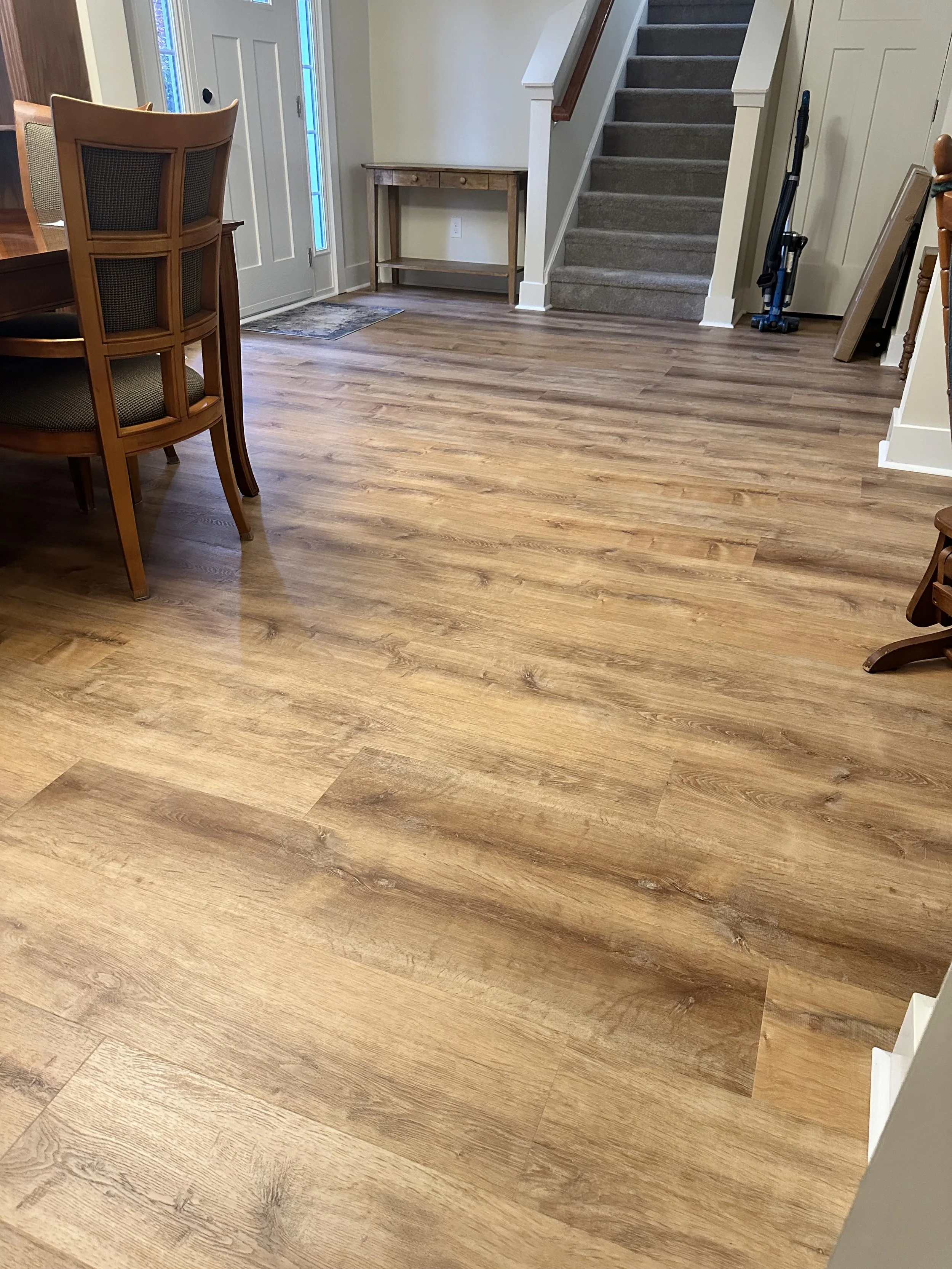 Living or dining room with wood-look flooring, a part of a wooden dining table with chairs, a staircase with carpeted steps, a small wooden table, and a doorway with glass panels.