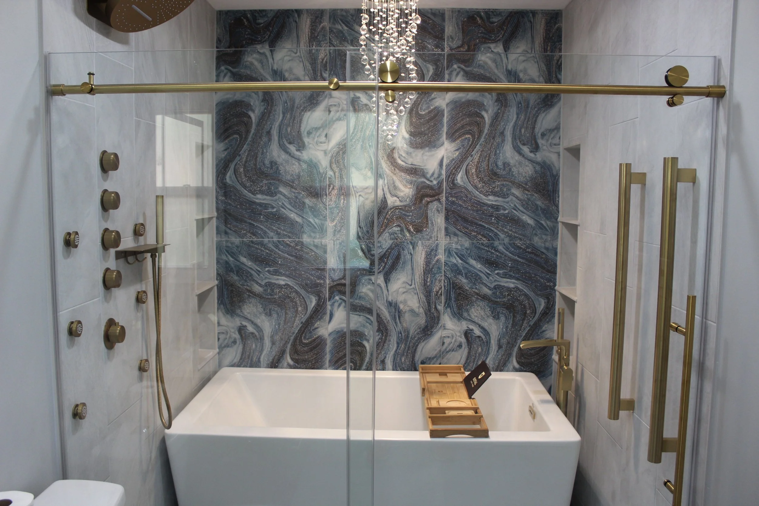 Modern bathroom with a freestanding bathtub, gold fixtures, a marble accent wall, a shower with multiple shower controls, glass door, built-in shelves, and a chandelier