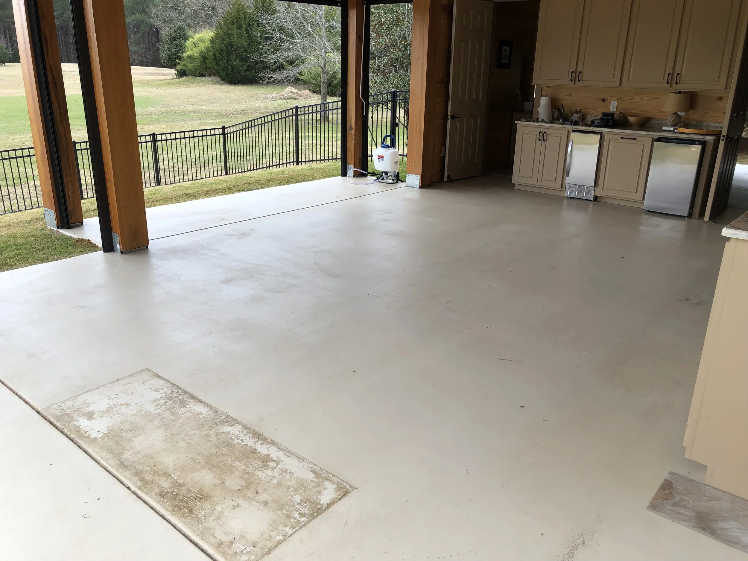 Empty garage with open doors leading to an outdoor grassy yard, with kitchen cabinets and appliances on the right side and a concrete floor.