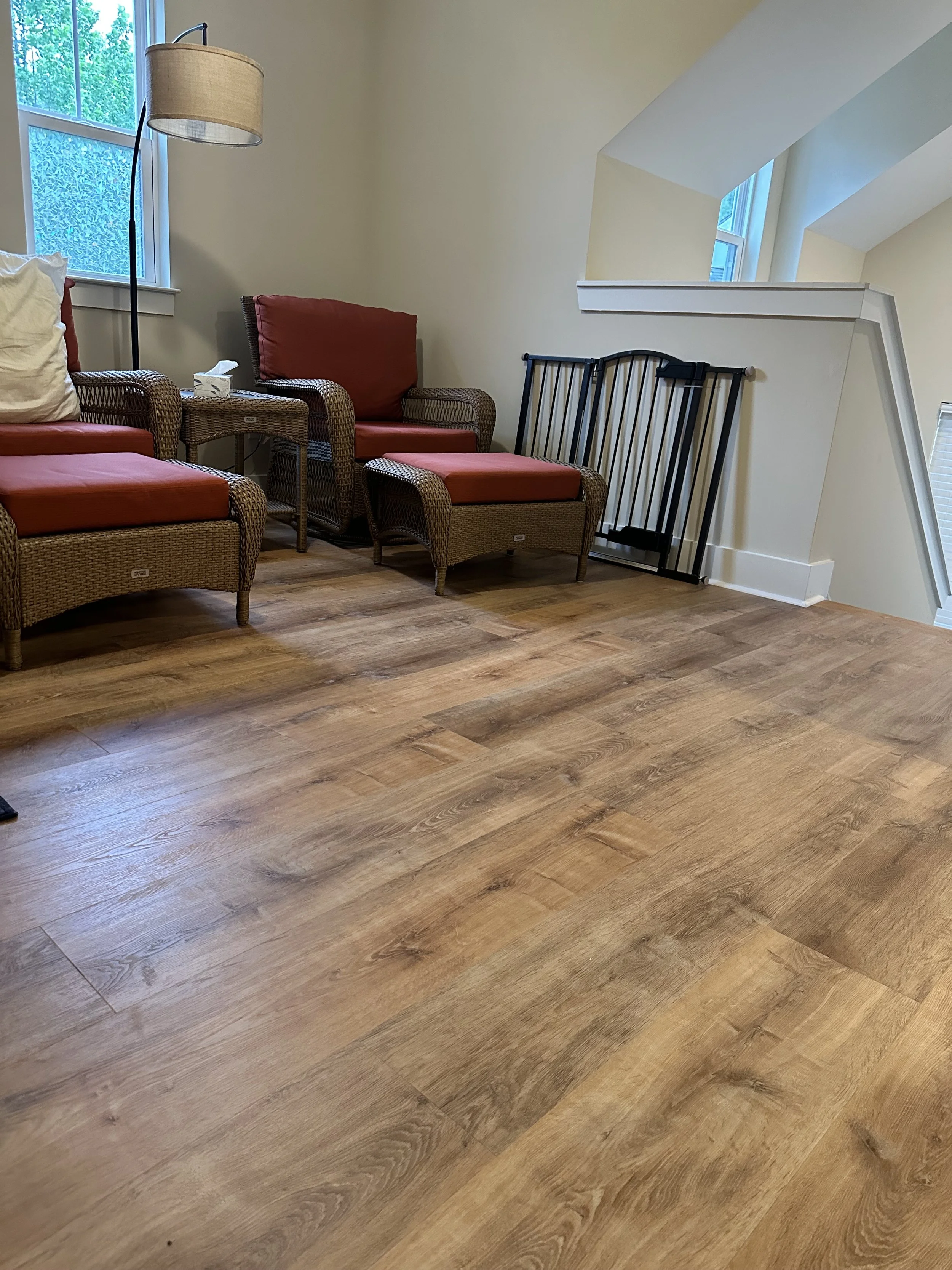 Living room with wooden floor, wicker furniture with red cushions, a floor lamp, and windows showing outside greenery.