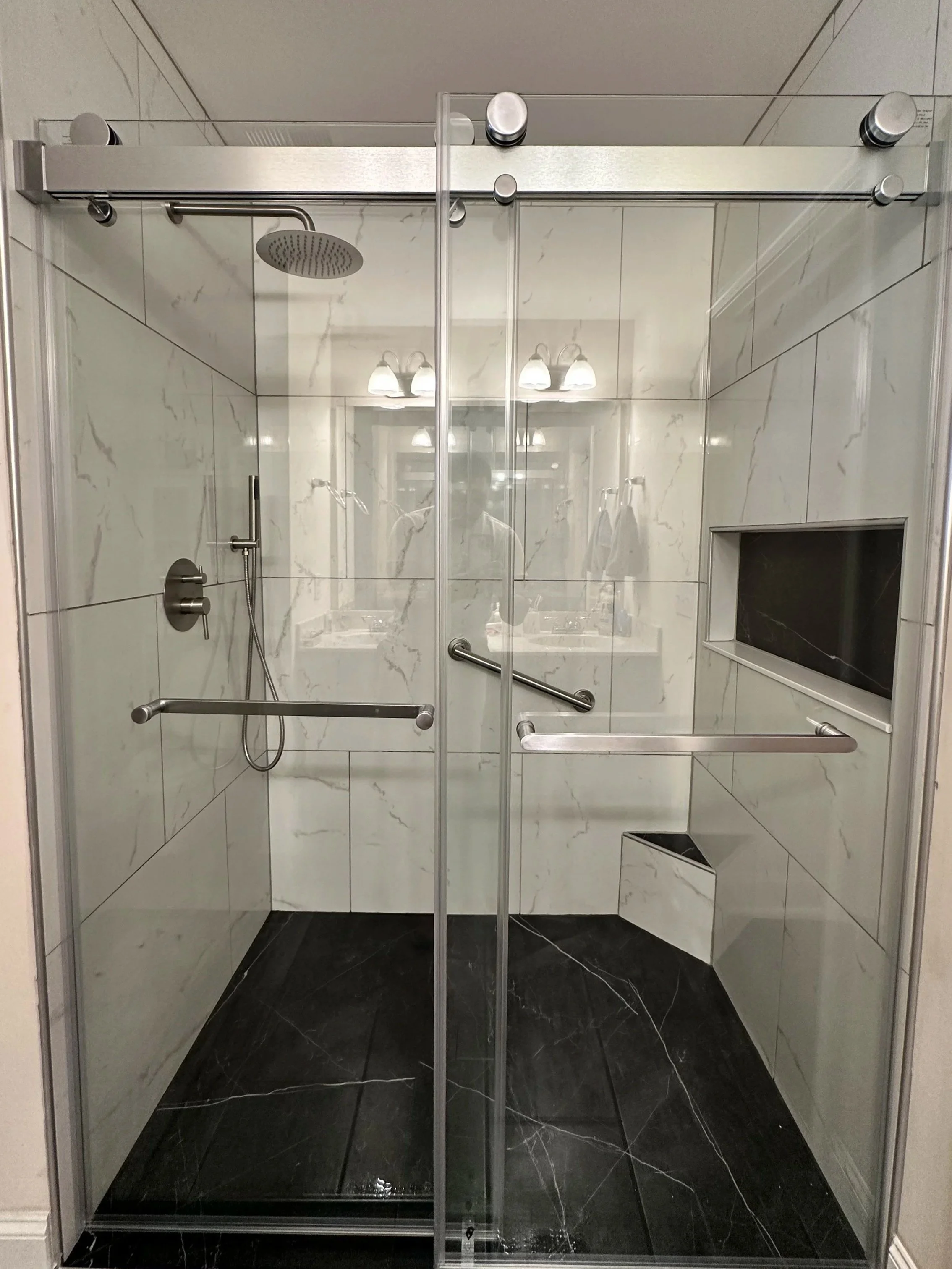 Walk-in shower with glass doors, a showerhead, a handheld shower, a built-in niche for toiletries, and black marble floor tiles.