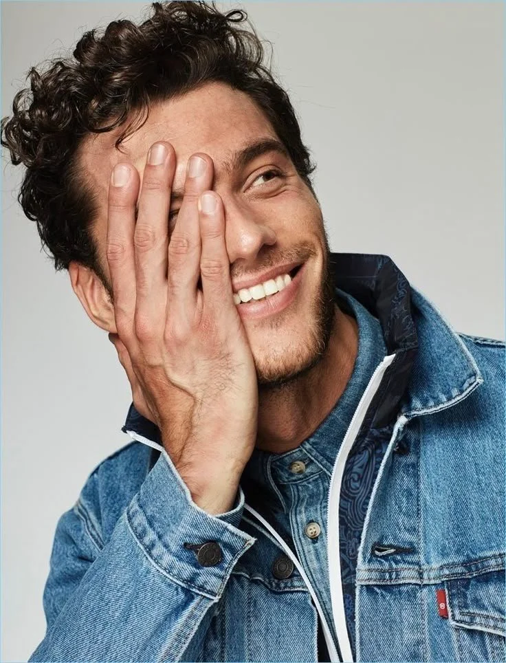 A young man with curly brown hair and a beard smiling, covering one eye with his hand, dressed in a blue denim jacket and shirt, against a plain background.