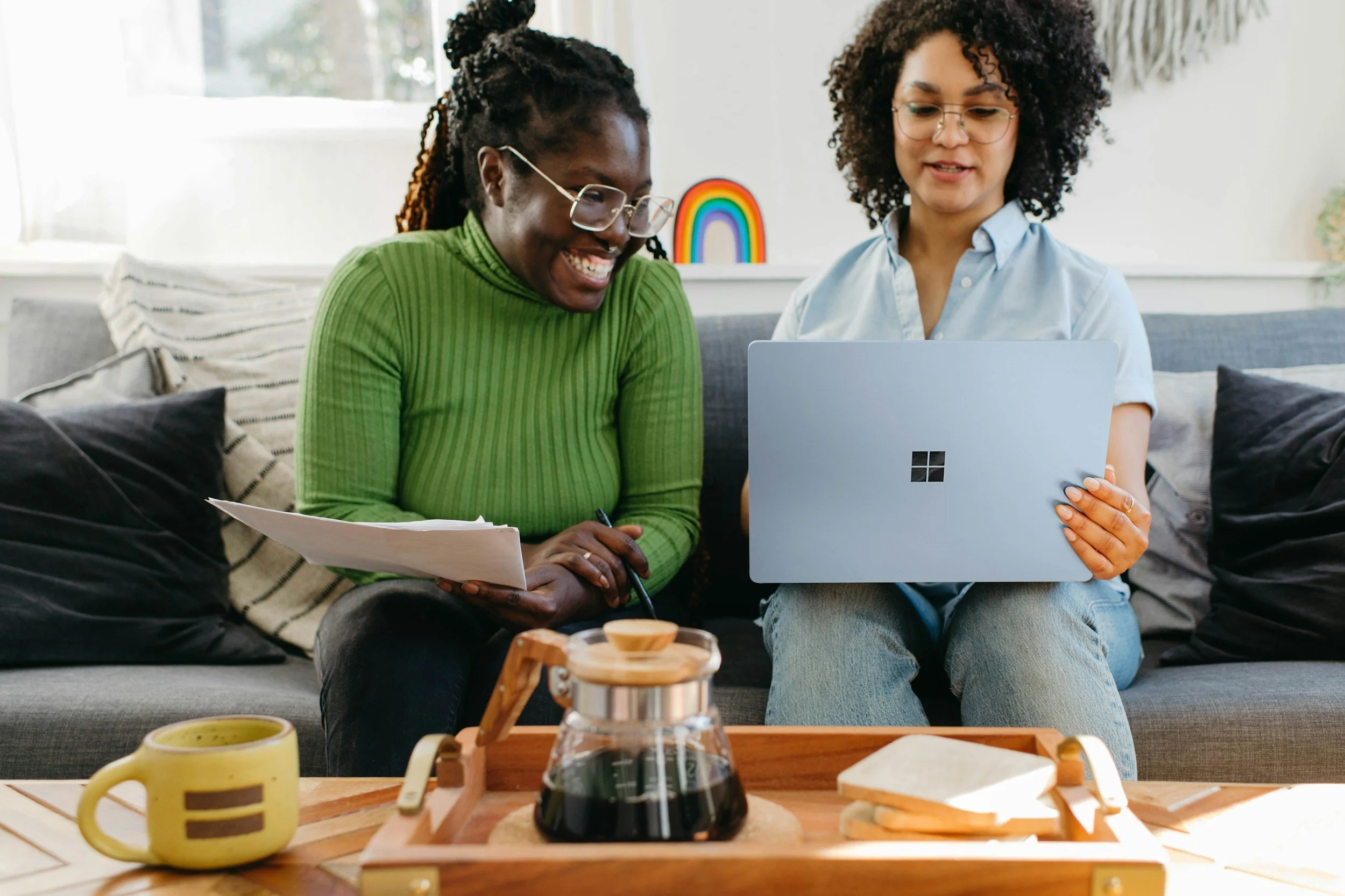 Two women sitting on a gray sofa in a bright living room, looking at a laptop together. One woman is wearing a green sweater and glasses and holding papers, while the other woman is wearing a light blue shirt and glasses. There are cushions, a tray with a coffee pot, a mug, and slices of bread on the table in front of them.