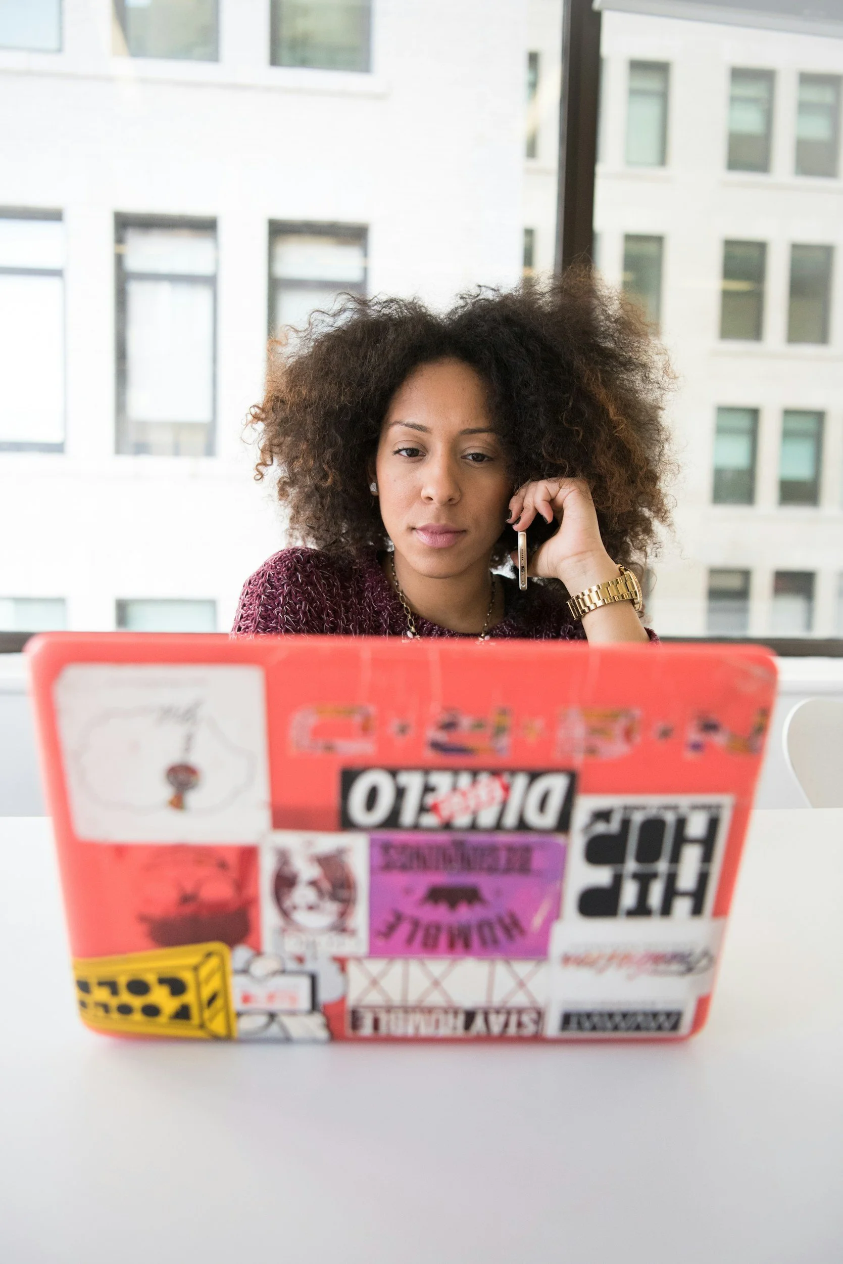 A young woman with curly hair and a gold watch talking on her phone, working on a laptop covered with colorful stickers, sitting at a table near a window in an office or cafe with buildings visible outside.