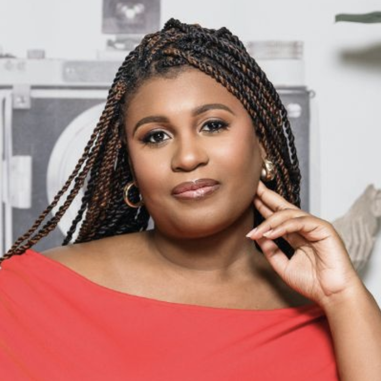 Portrait of a confident woman with braided hair and gold hoop earrings, wearing a coral top, standing in front of a laundry washing machine and dryer in a laundry room.