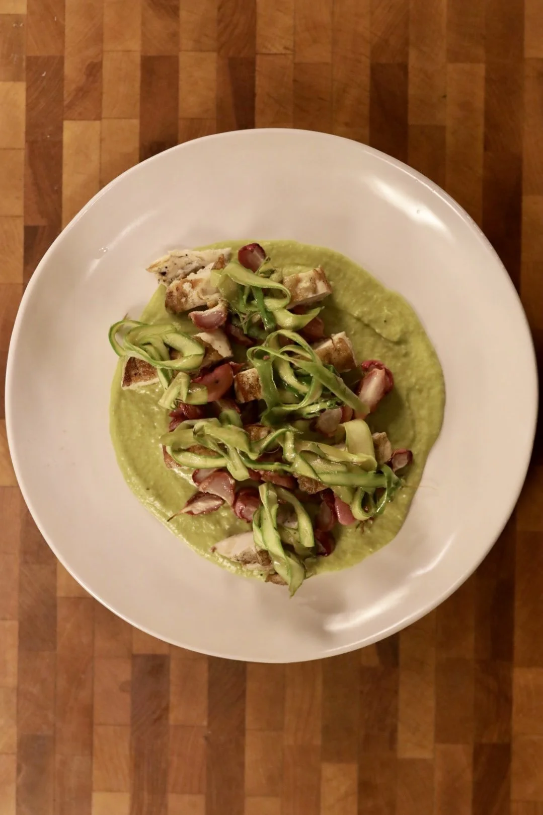 A dish with green puree topped with shredded zucchini and chunks of grilled chicken, served on a white plate on a wooden surface.