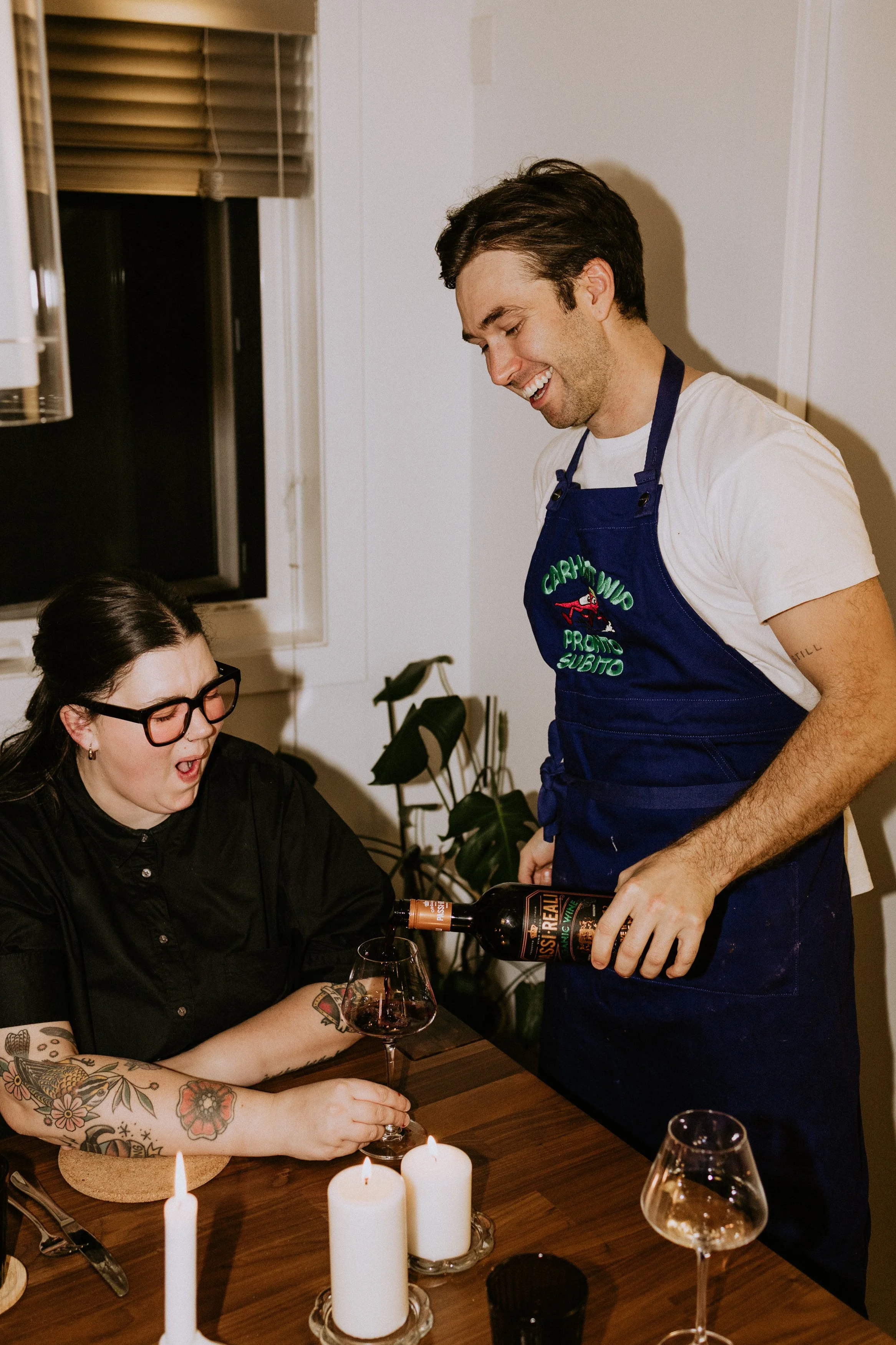 A man in a white t-shirt and blue apron pouring red wine into a woman's glass at a dinner table, with lit candles and wine glasses present.