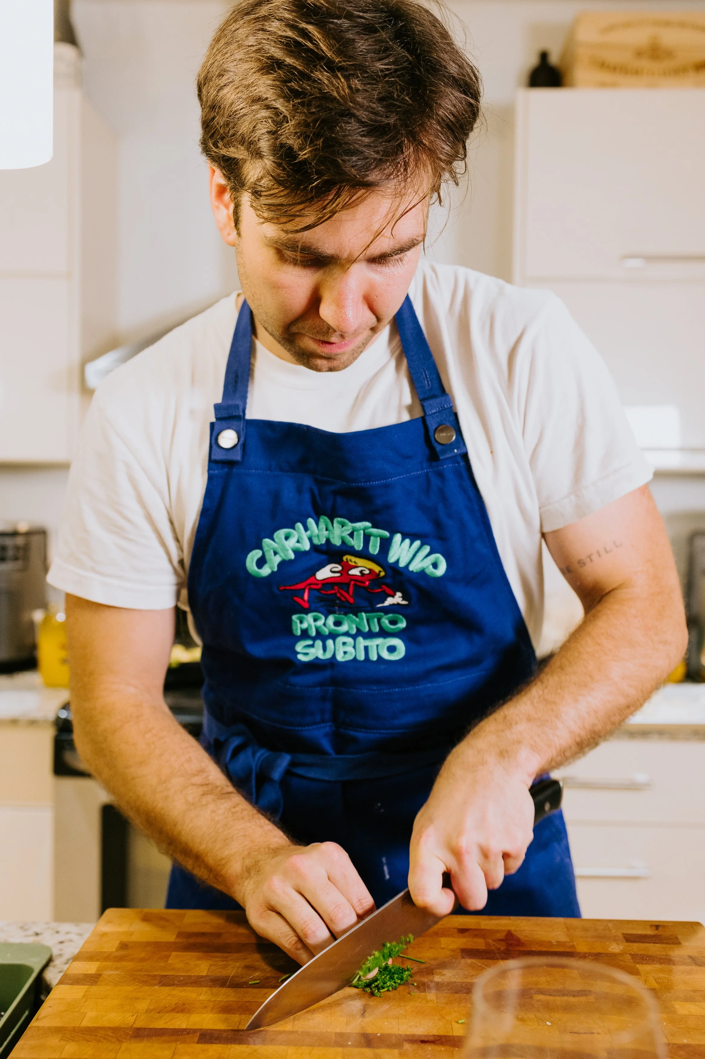 A man wearing a blue apron slicing parsley on a wooden cutting board in a kitchen.