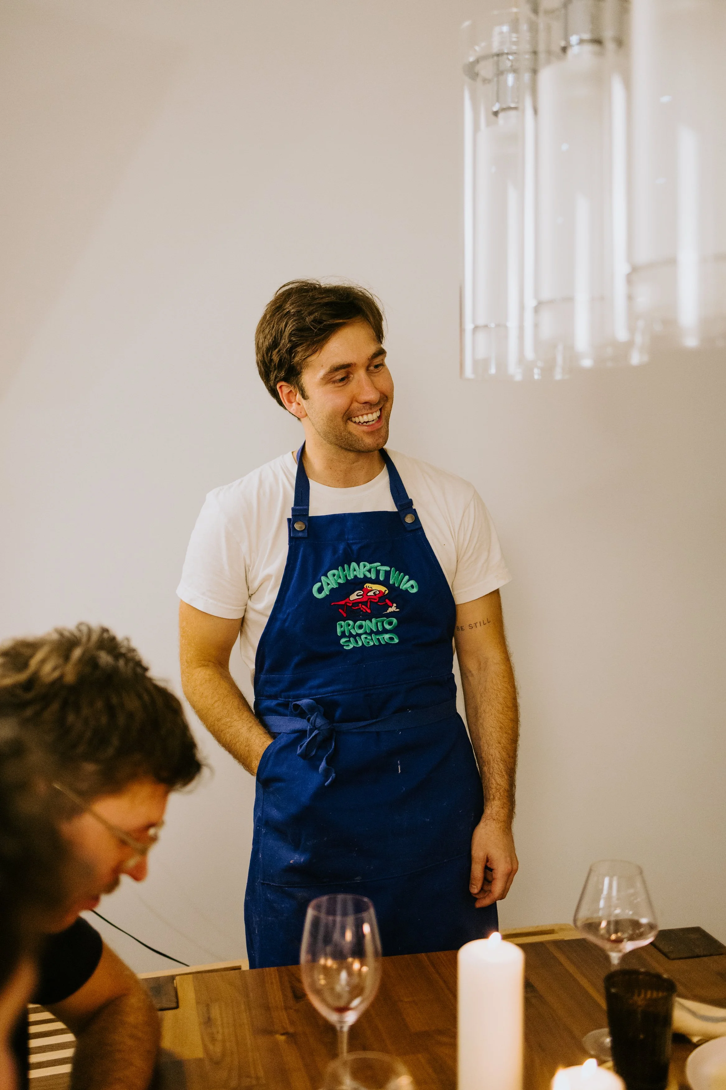 A man wearing a Carhartt work apron and white t-shirt, smiling, at a dinner table with glasses and candles.