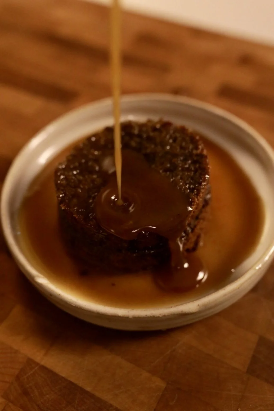 A piece of meat with sauce being poured over it in a white bowl on a wooden surface.