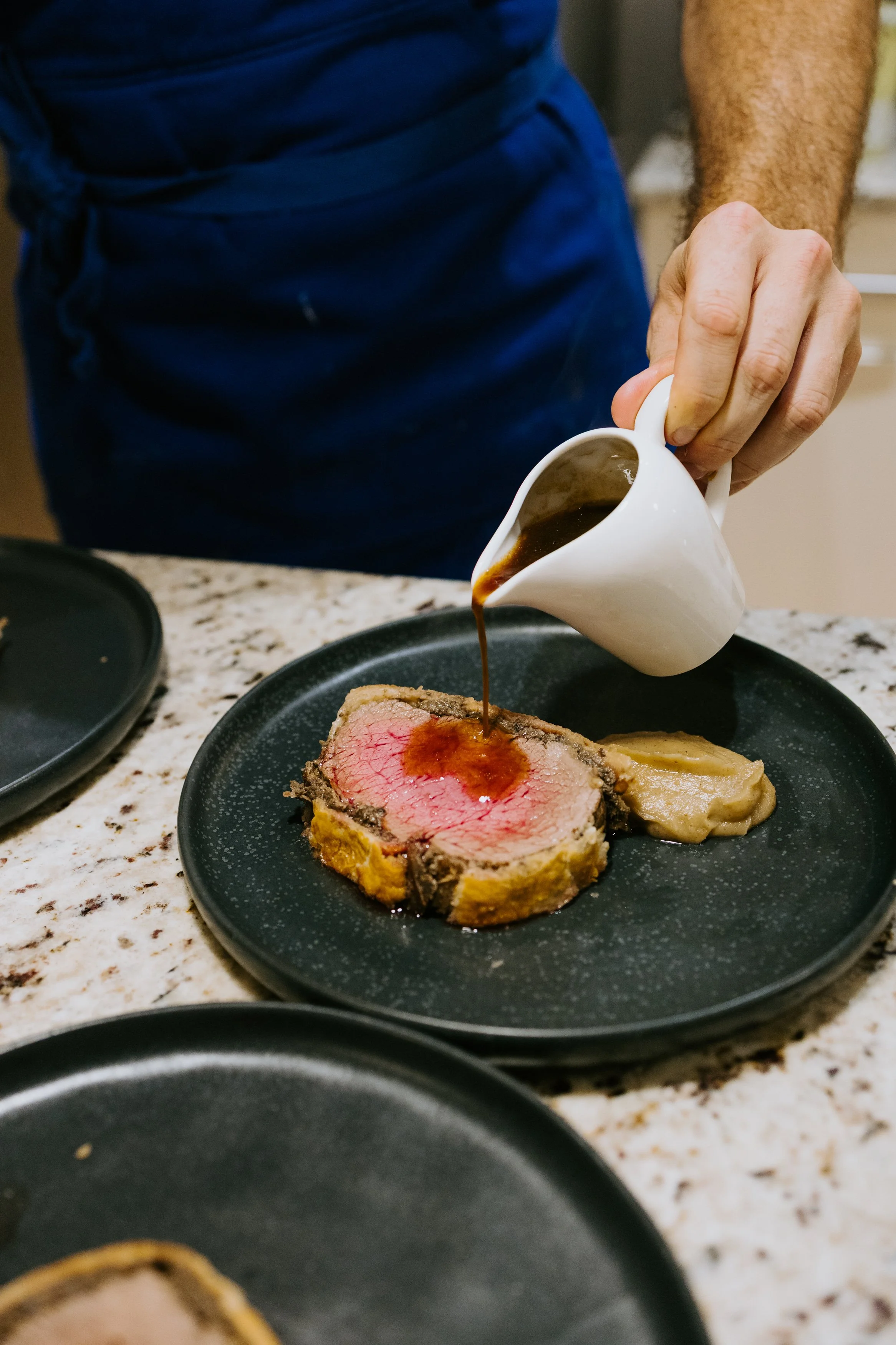 Person pouring brown gravy on medium-rare beef Wellington with mashed potatoes on black plate.